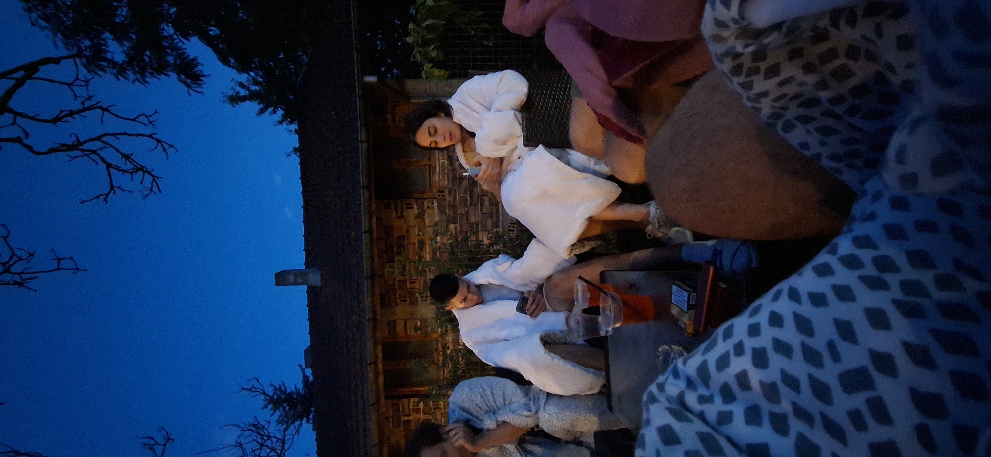 The image depicts a group of people, some wearing white bathrobes, gathered outdoors at dusk. They are seated around a table, with drinks and a pack of cigarettes visible. In the background, a rustic brick building with a tiled roof is present, suggesting a rural or suburban setting. The overall atmosphere appears relaxed and social. The specific location is identified as Lobosh, Bulgaria, though the exact nature of the gathering and the building are not explicitly discernible from the visual information provided.