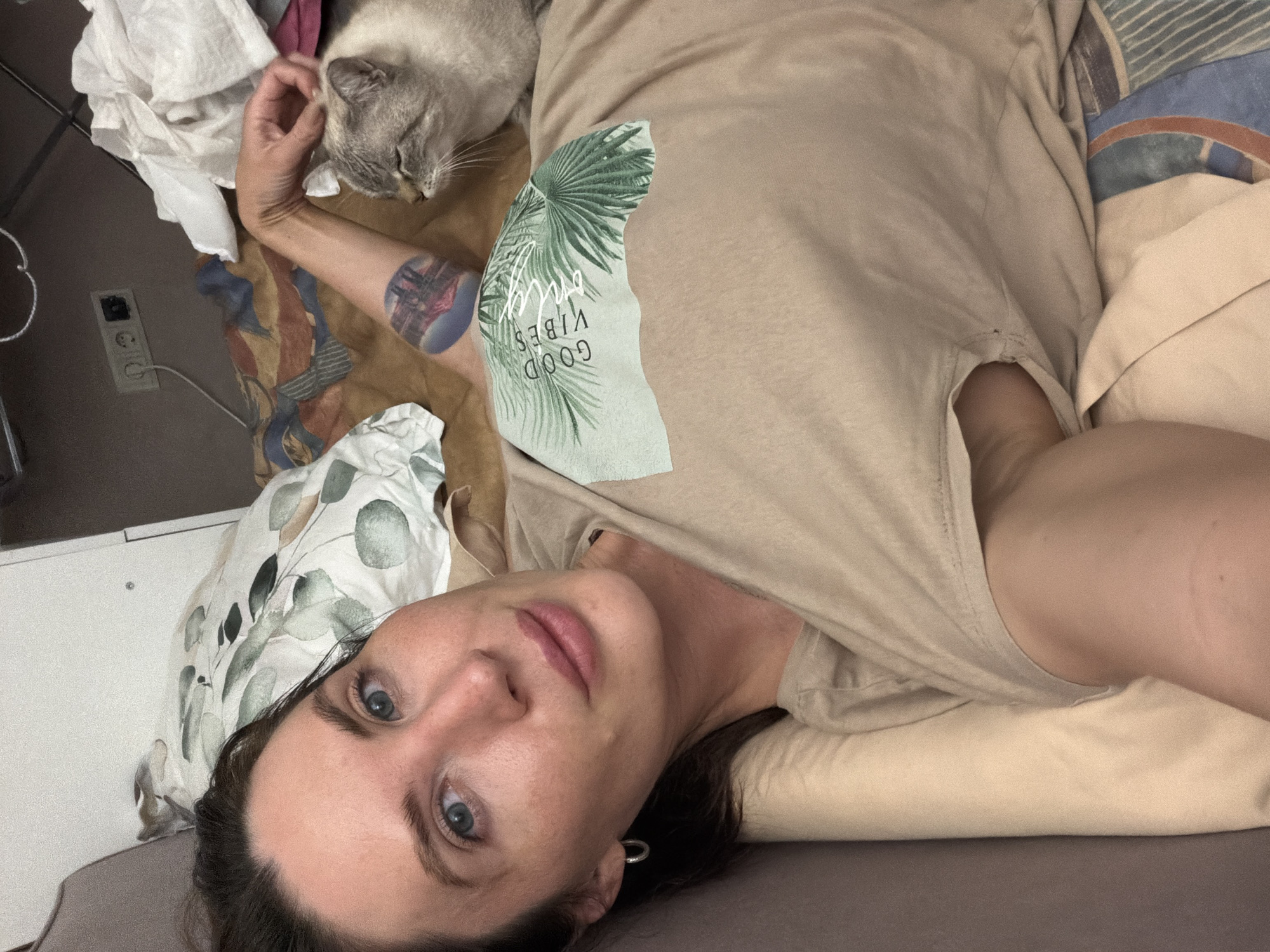 A woman with fair skin, blue eyes, and brown hair is lying down, facing the camera. She is wearing a light brown t-shirt with a palm leaf graphic and the words "GOOD VIBES". A cat with gray and white fur is lying next to her, and she is petting it. The background appears to be a bedroom with pillows and blankets. There is an electrical outlet visible on the wall. The scene suggests a relaxed, intimate moment shared between the woman and her pet. The location is identified as Perm, Russia.