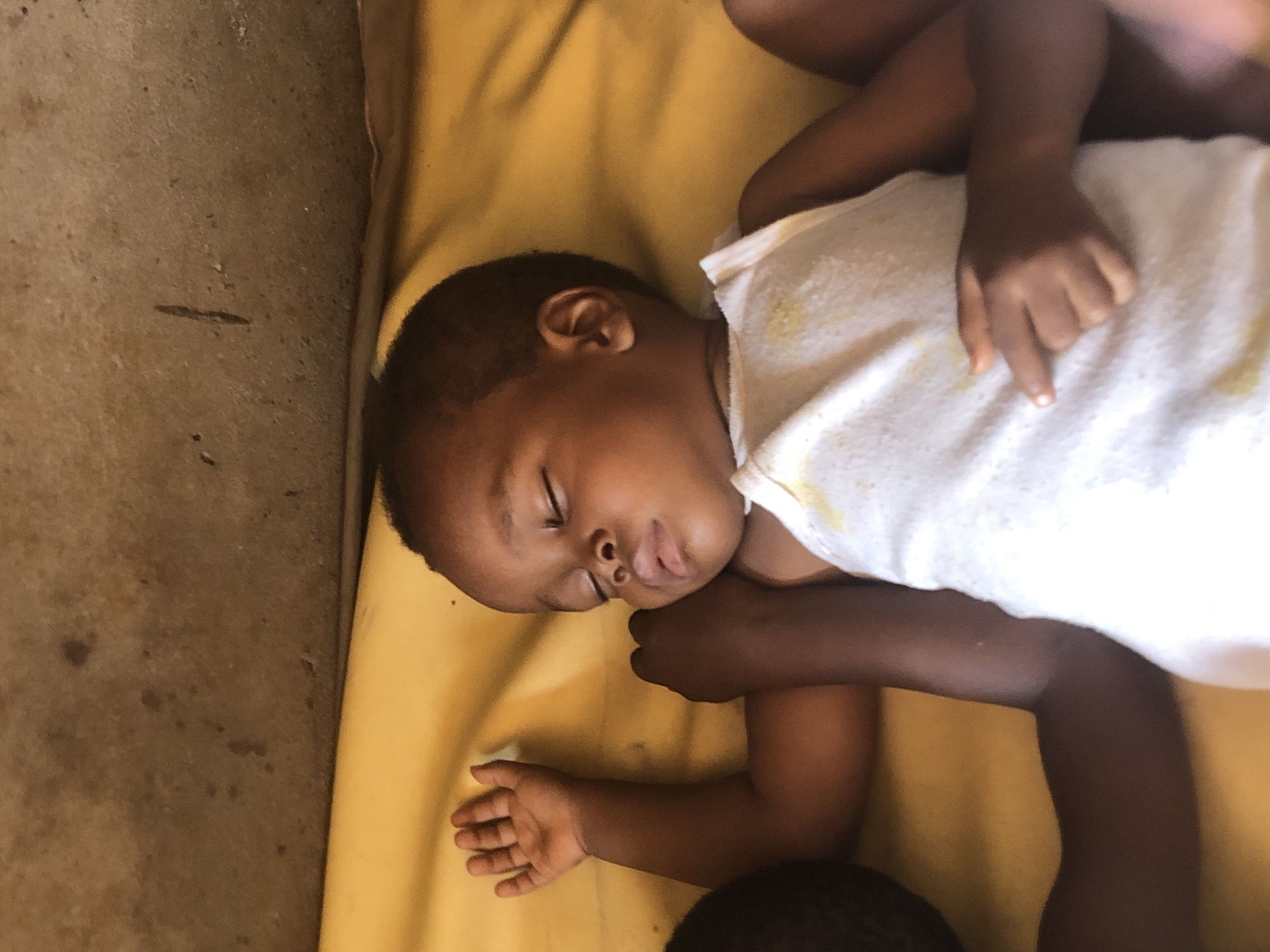 A young child, appearing to be asleep, is lying on a yellow surface, likely a mat or blanket. The child is wearing a white tank top with faint yellow marks. The child's right arm is extended with the hand open, fingers slightly curled. The child's head is turned to the right, with eyes closed and mouth slightly open. Another person's arm is visible under the child's head and arm. In the background, a textured, brown wall or ceiling is visible. The overall scene suggests an indoor or covered outdoor environment. This scene is located in Ukwu-Abwa, Nigeria.