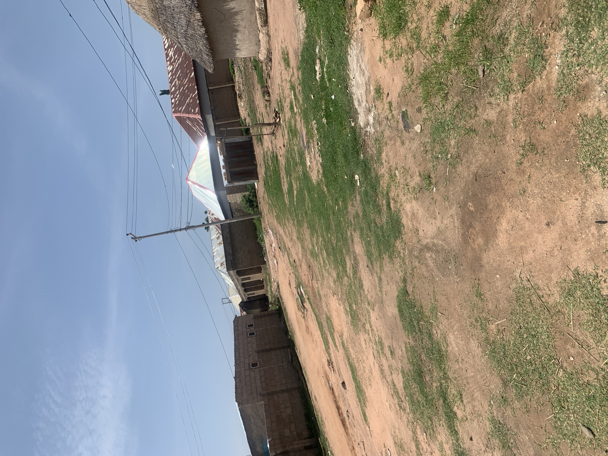 The image depicts a street scene in Jauro Yinu, Nigeria, under a clear blue sky with sparse white clouds. The ground is a mix of reddish-brown dirt and sparse green grass. Several structures are visible, including buildings constructed with brick and concrete blocks, some with corrugated metal roofs and others with thatched roofs. A prominent feature is a covered, multi-sided structure with a metallic roof, likely a shelter or market stall, situated in the mid-ground. Electrical wires run overhead, supported by a utility pole. The environment suggests a rural or semi-urban setting, with evidence of ongoing construction or unpaved roads. No individuals are clearly visible in the frame.
