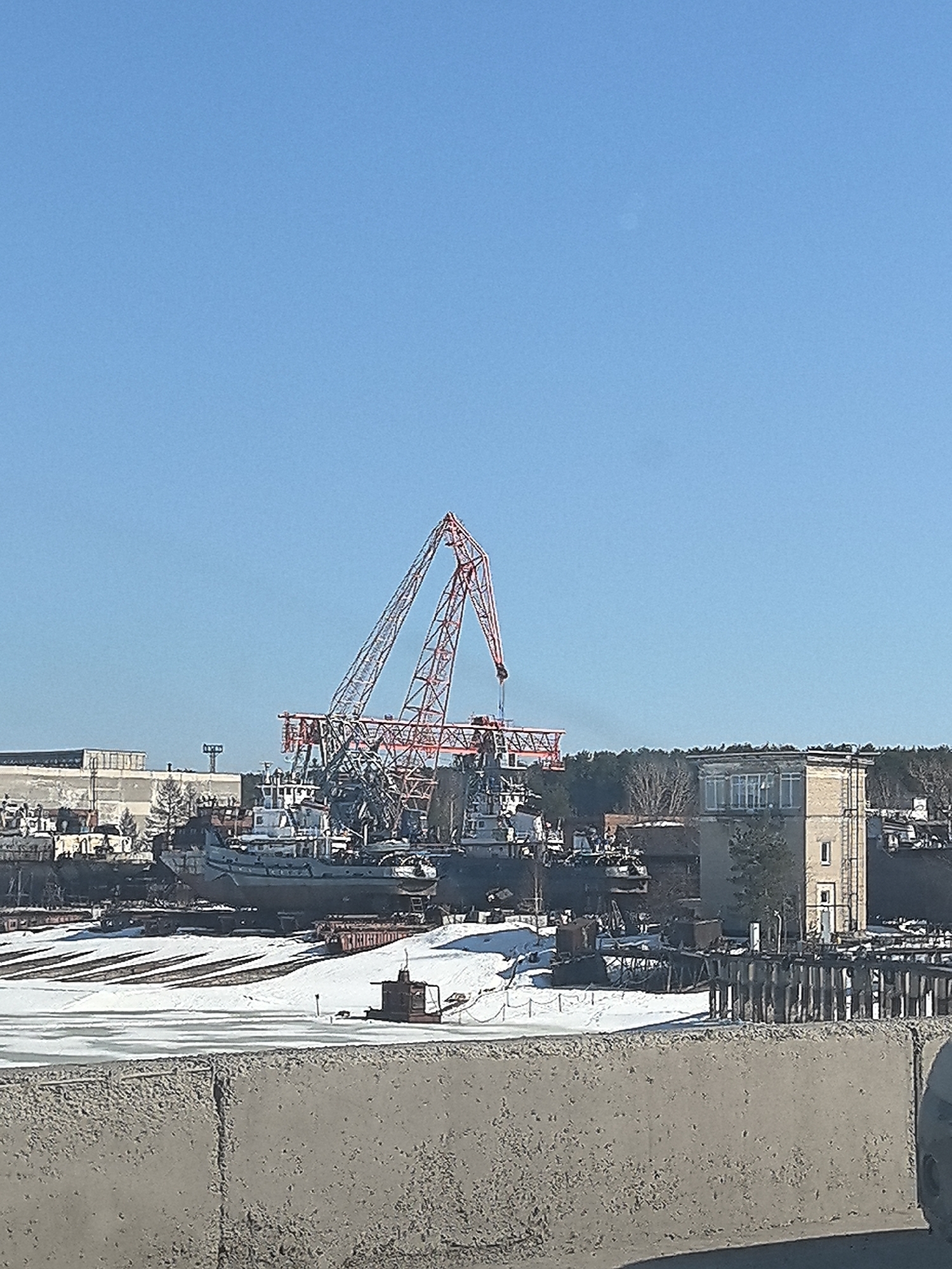 The image shows a shipyard or repair facility. The scene features a large, red gantry crane dominating the center of the frame. There are several ships of various sizes docked, some possibly under repair or maintenance. The ground is covered in snow, suggesting a cold climate. A concrete barrier is in the foreground, obscuring part of the view. In the background, there are industrial buildings and sparse vegetation. Based on the request, the location is in the city of Chaikovsky, Russia.