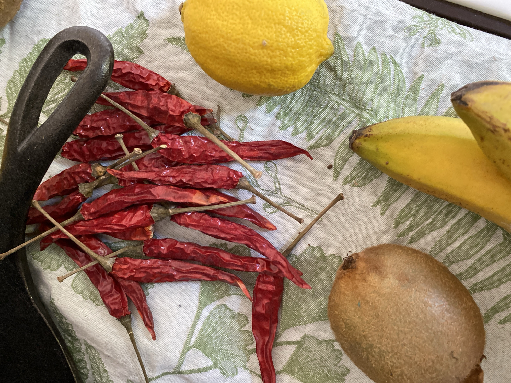 A still life arrangement of dried red chili peppers, a lemon, a kiwi, and a banana is laid out on a white cloth with a green fern pattern. A black cast iron pot handle is visible in the upper left corner of the frame, partially obscuring a cluster of chili peppers. The objects are arranged on a flat surface. The scene is indoors. There are no people or actions depicted. The location is not specified within the image.