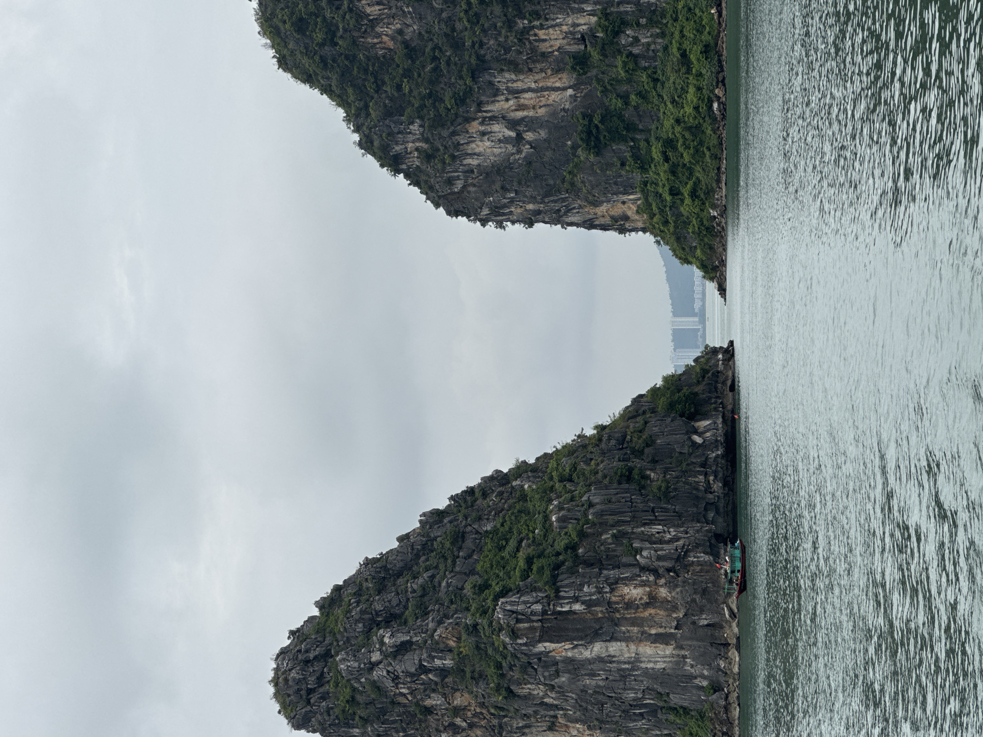 The image depicts a serene seascape with two imposing limestone karst islands flanking a channel of water. The islands, lush with green vegetation clinging to their steep, rocky faces, rise dramatically from the sea. The sky above is overcast with a pale gray hue, suggesting a diffused light. In the distance, beyond the channel, a modern city skyline with tall buildings is visible, indicating the presence of urban development near this natural wonder. On the water, a small, green boat with a red trim is navigating through the calm sea, adding a touch of human activity to the otherwise natural landscape. The water itself exhibits gentle ripples, reflecting the muted tones of the sky and islands. The scene captures a juxtaposition of natural beauty and human presence, characteristic of many coastal areas in Tuan Chau Ward, Vietnam, which is renowned for its limestone karsts and bays.