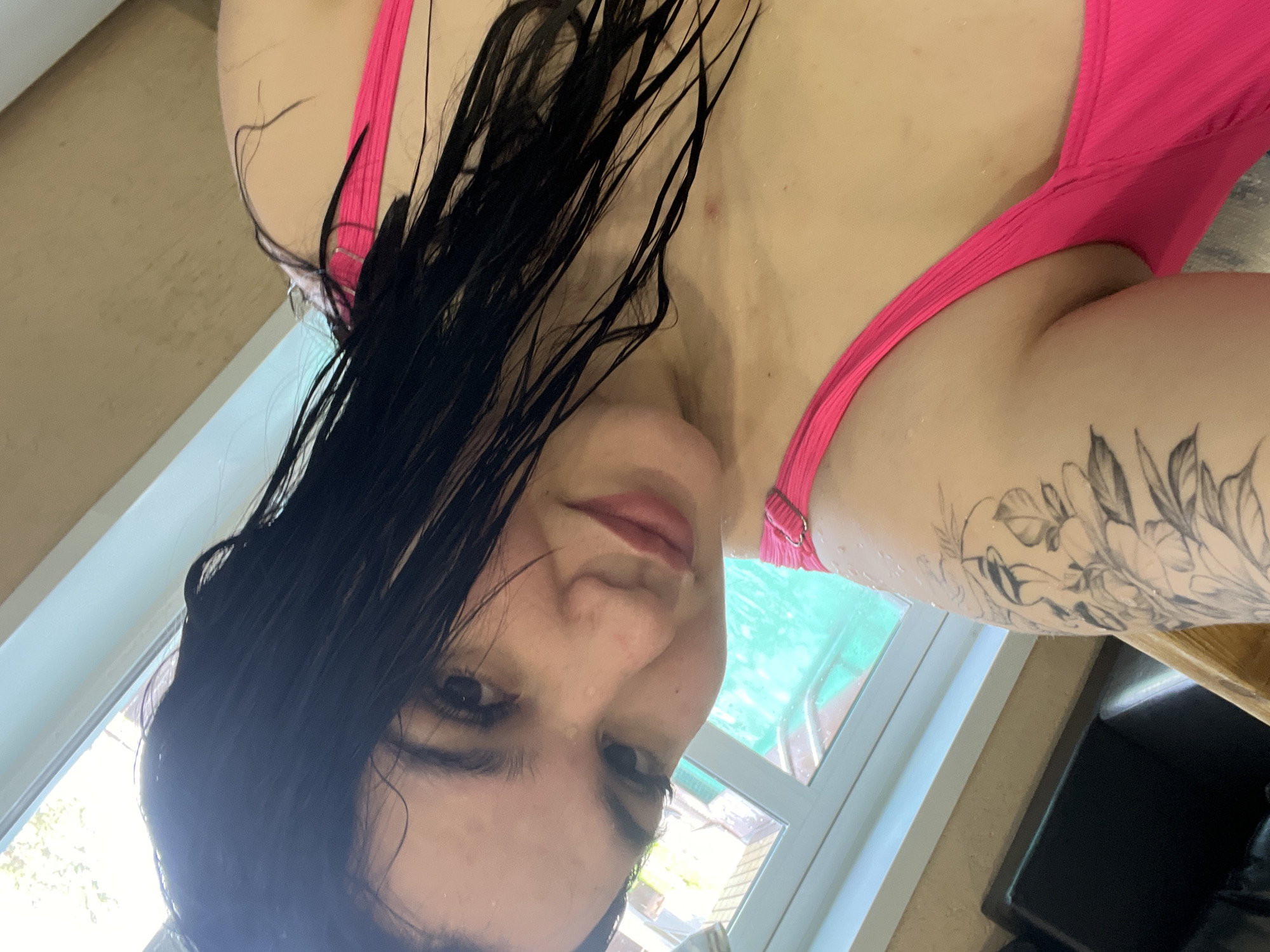 A woman with wet dark hair and a pink bathing suit is in front of a window. The location is possibly indoors, with a view of a swimming pool visible through the window. She has a tattoo on her left arm. The location is Chaikovsky, Russia.