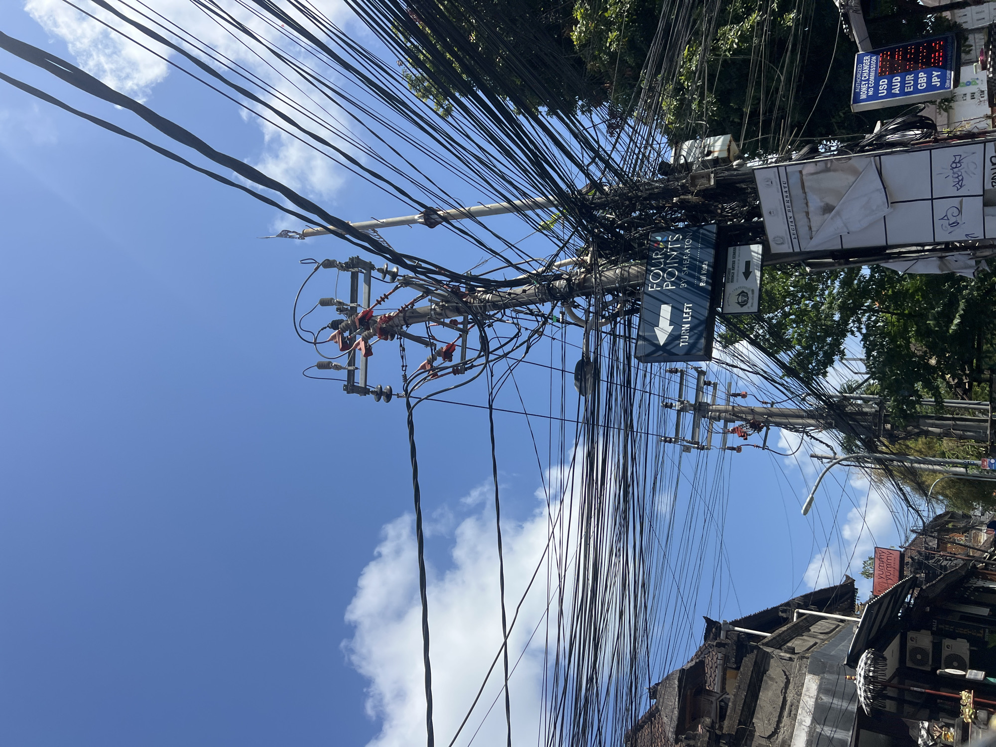 This is a low-angle, wide shot of a street scene in Kuta, Indonesia. A cluster of utility poles with a dense entanglement of black electrical wires dominates the center of the frame, against a clear blue sky with scattered white clouds. The poles are equipped with transformers and insulators. A directional sign for "Four Points by Sheraton" and a "Turn Left" arrow is visible on one of the poles. To the left of the utility poles, a traditional Balinese building with a tiled roof and intricate carvings is partially visible. A street umbrella is set up in front of a storefront. To the right of the utility poles, dense green foliage is seen. A sign for an "Authorized Money Changer" displaying exchange rates for USD, AUD, EUR, GBP, and JPY is mounted on another pole in the foreground. The scene appears to be during daytime.