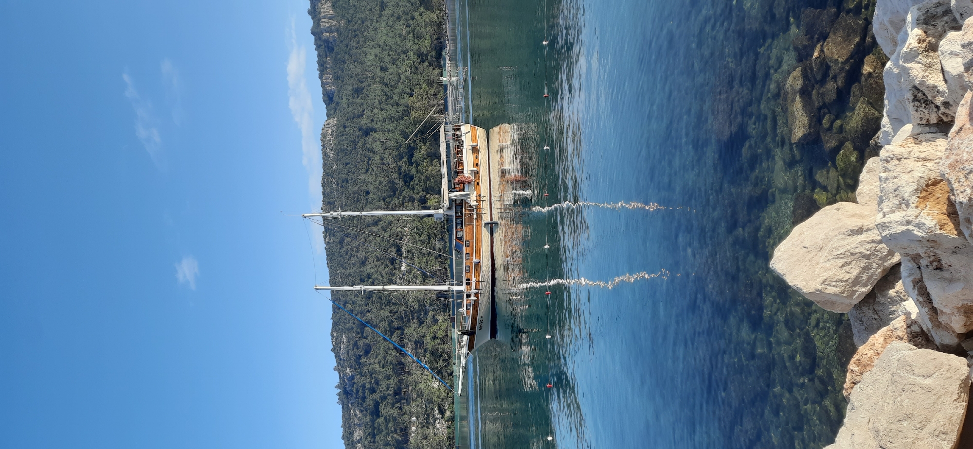 The image captures a boat scene in what appears to be the city of Kemer, Turkey, given the coastal environment and Mediterranean vegetation. A multi-masted sailing boat is positioned near the coastline. The water is clear, revealing underwater rocks near the shore. A rocky outcrop is visible along the edge of the water. The landscape includes lush green hills in the background under a clear blue sky with scattered clouds.