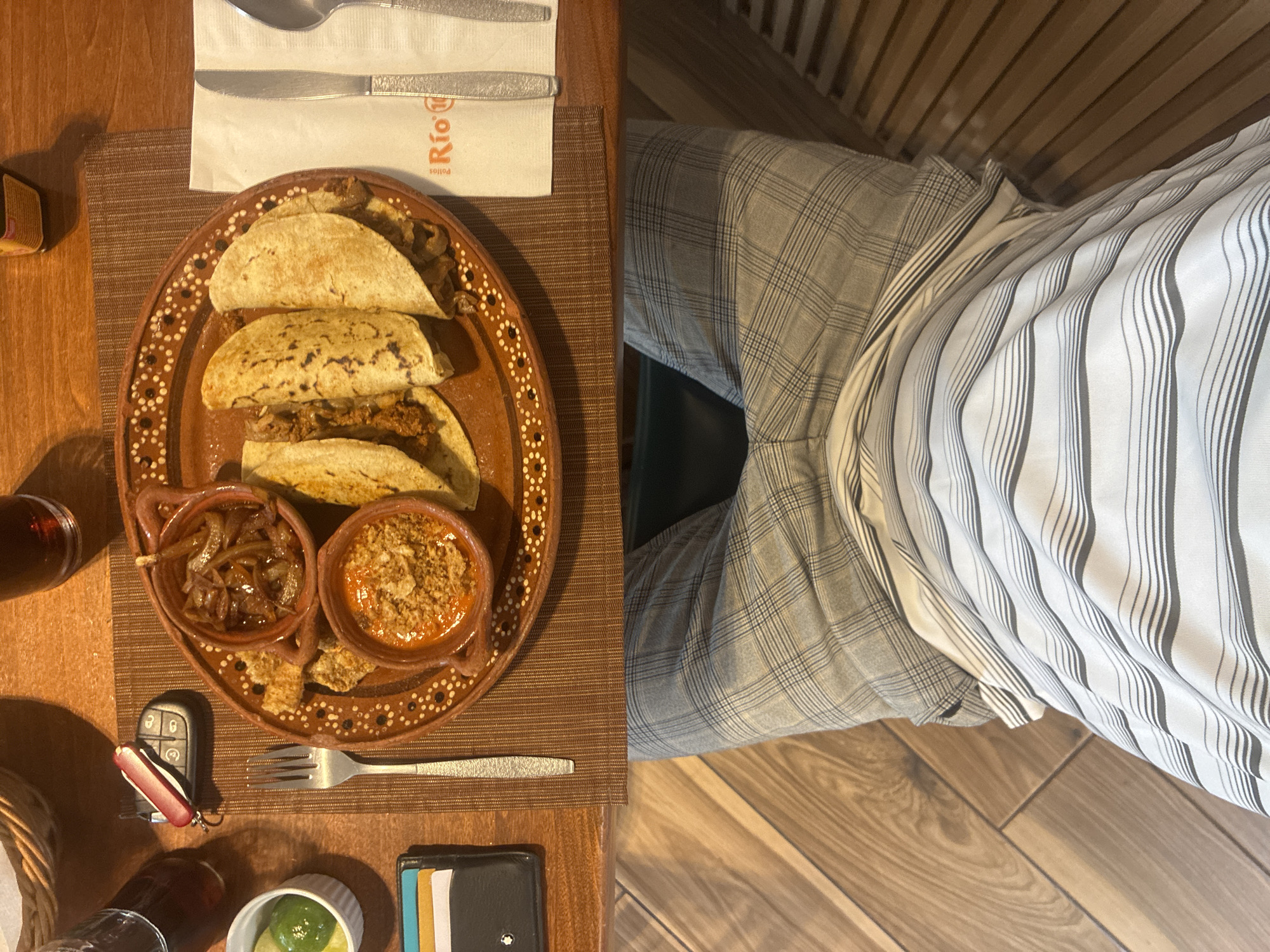 The image shows a table with a plate of tacos, two small bowls of toppings, silverware, a napkin, a car key, a wallet, and lime wedges. The person sitting at the table is wearing plaid pants and a striped shirt. The location is likely a restaurant in Cuautitlán Izcalli, Mexico, based on the Spanish writing on the napkin.