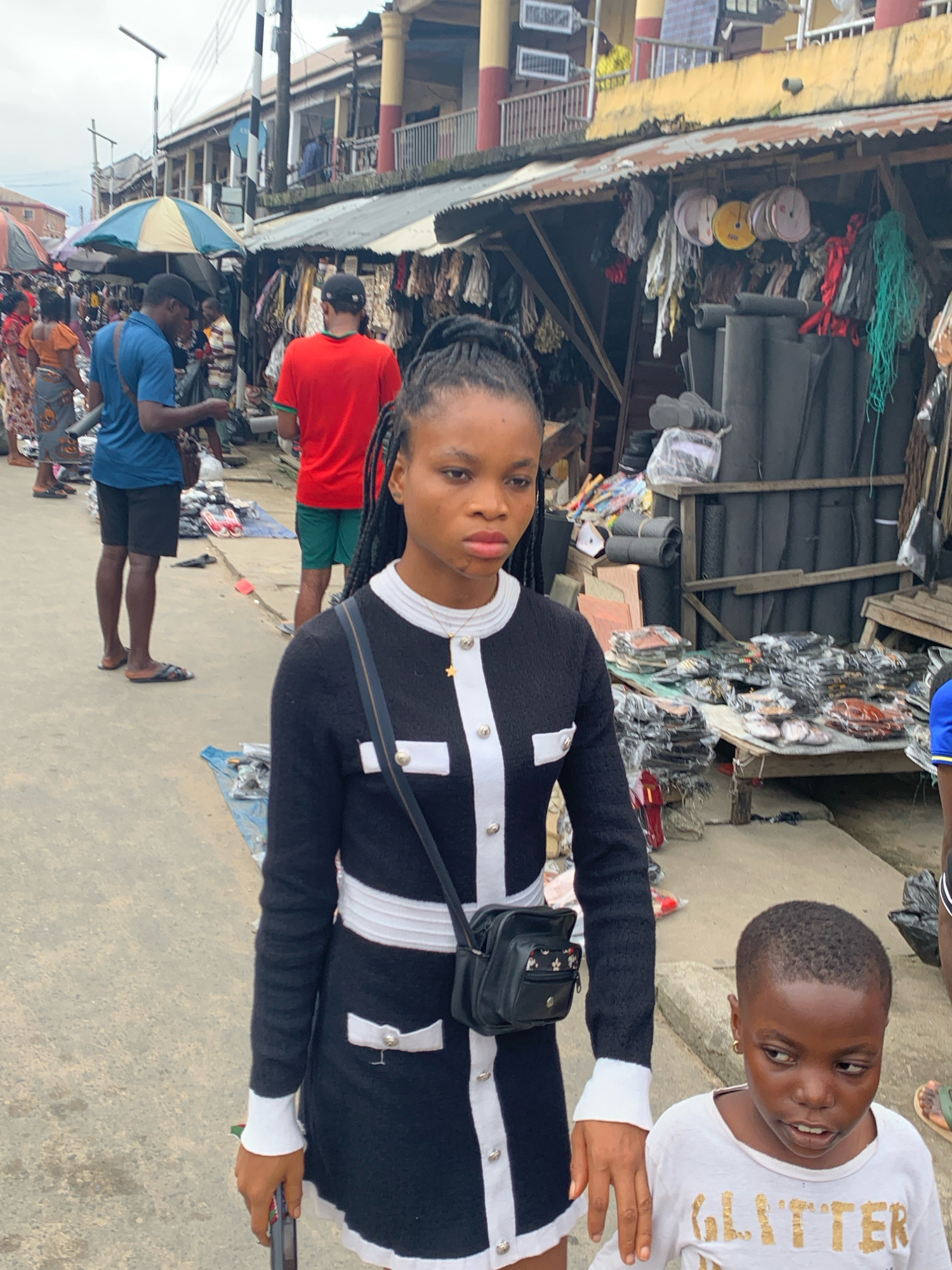 This is a photograph taken in Aba, Nigeria, depicting a bustling market scene. In the foreground, a young Black woman with braided hair and a black and white patterned dress walks towards the camera, carrying a small black bag over her shoulder. Beside her, a young Black child in a white t-shirt with "GLITTER" printed on it looks to the side. The background shows a busy marketplace with stalls selling various goods, including fabrics, rolls of material, and what appear to be shoes or sandals. Several other individuals, including men in blue and red shirts and women in colorful attire, are visible in the market. The environment appears to be an outdoor urban setting with buildings and overhead wires visible in the distance. The ground is paved, and there are various items laid out on tarps.