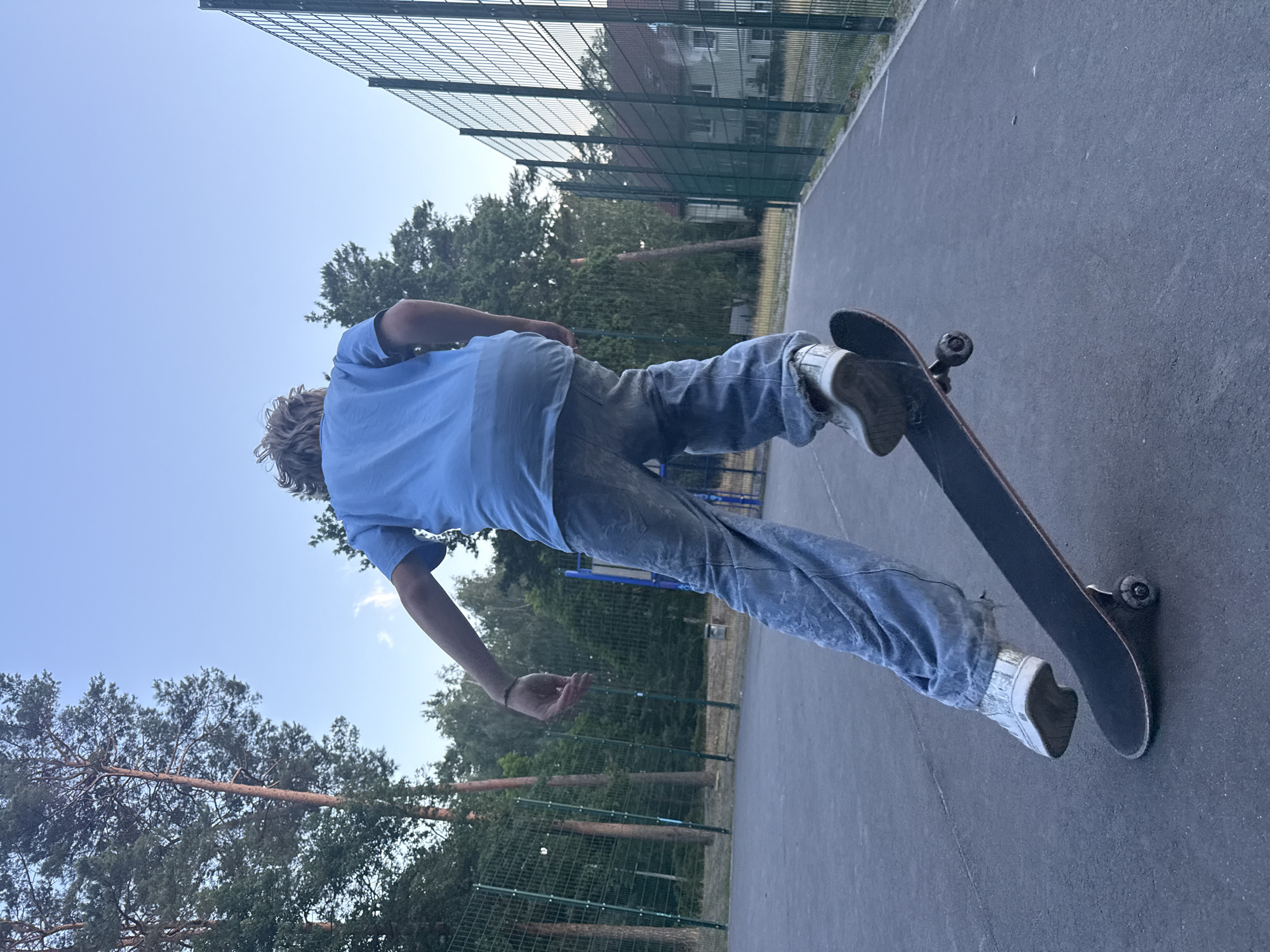 A person is skateboarding on an asphalt court in Eberswalde, Germany. The person is seen from behind, wearing a light blue t-shirt and loose-fitting denim jeans. They are on a black skateboard with visible wheels and trucks. The court is surrounded by a green chain-link fence, behind which are trees and buildings. The sky above is a clear blue with a few wispy clouds. The lighting suggests it is late afternoon or early evening. The perspective is low, looking up at the skateboarder.