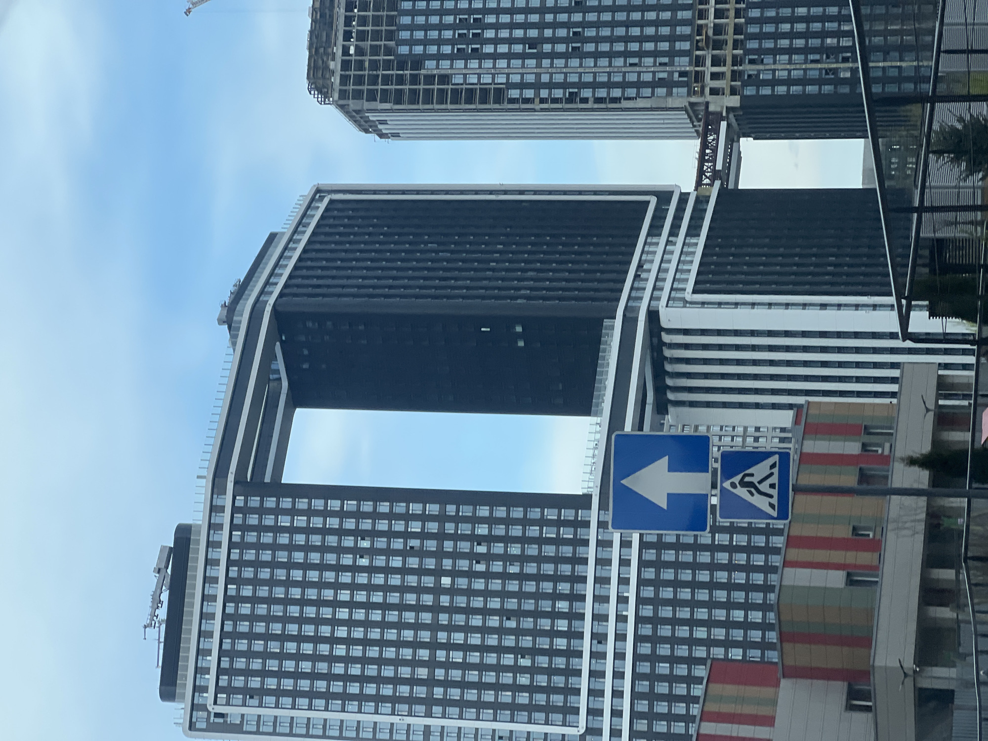 The image depicts a modern architectural scene in Khimki, Russia. The focal point is a high-rise building characterized by a large rectangular void in its upper section. The building features a grid-like facade with numerous windows. Another high-rise structure, in a state of construction, is visible in the background, revealing its unfinished framework. In the foreground, a street sign with a white arrow pointing left and a pedestrian crossing sign are prominently displayed. The sky is partly cloudy. The overall environment suggests an urban setting with contemporary architecture.