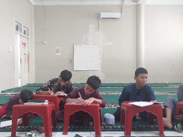 The scene depicts an interior view of a room, likely a religious school or mosque, located in Lhokseumawe, Indonesia. Four young males, appearing to be teenagers, are seated on a green patterned prayer rug that covers the floor. The walls are light-colored (off-white/beige) with visible wear, and a white air conditioning unit is mounted high on the right wall. A plain white board is affixed to the wall in the center. An open doorway is visible on the far left.
Each individual is positioned behind a small, red plastic stool with ornate cut-out designs, which serves as a low desk.
1. The male on the far left, wearing a maroon long-sleeved tunic and matching pants, is leaning forward over his stool, which holds a closed book. A small white and green bottle is on the floor near him.
2. The male second from the left, wearing a dark-patterned long-sleeved tunic, is actively writing on a sheet of white paper placed on his stool, holding a pen in his right hand.
3. The central male, also in a maroon long-sleeved tunic and matching pants, is similarly writing on a paper on his stool, holding a pen in his right hand.
4. The male on the far right, dressed in a dark blue long-sleeved tunic, is looking towards his paper on the stool and appears to be writing or preparing to write. A white skullcap is on the floor near his feet.
All individuals are focused on their writing or materials, with no direct interaction observed between them. The overall environment suggests a learning or study session.