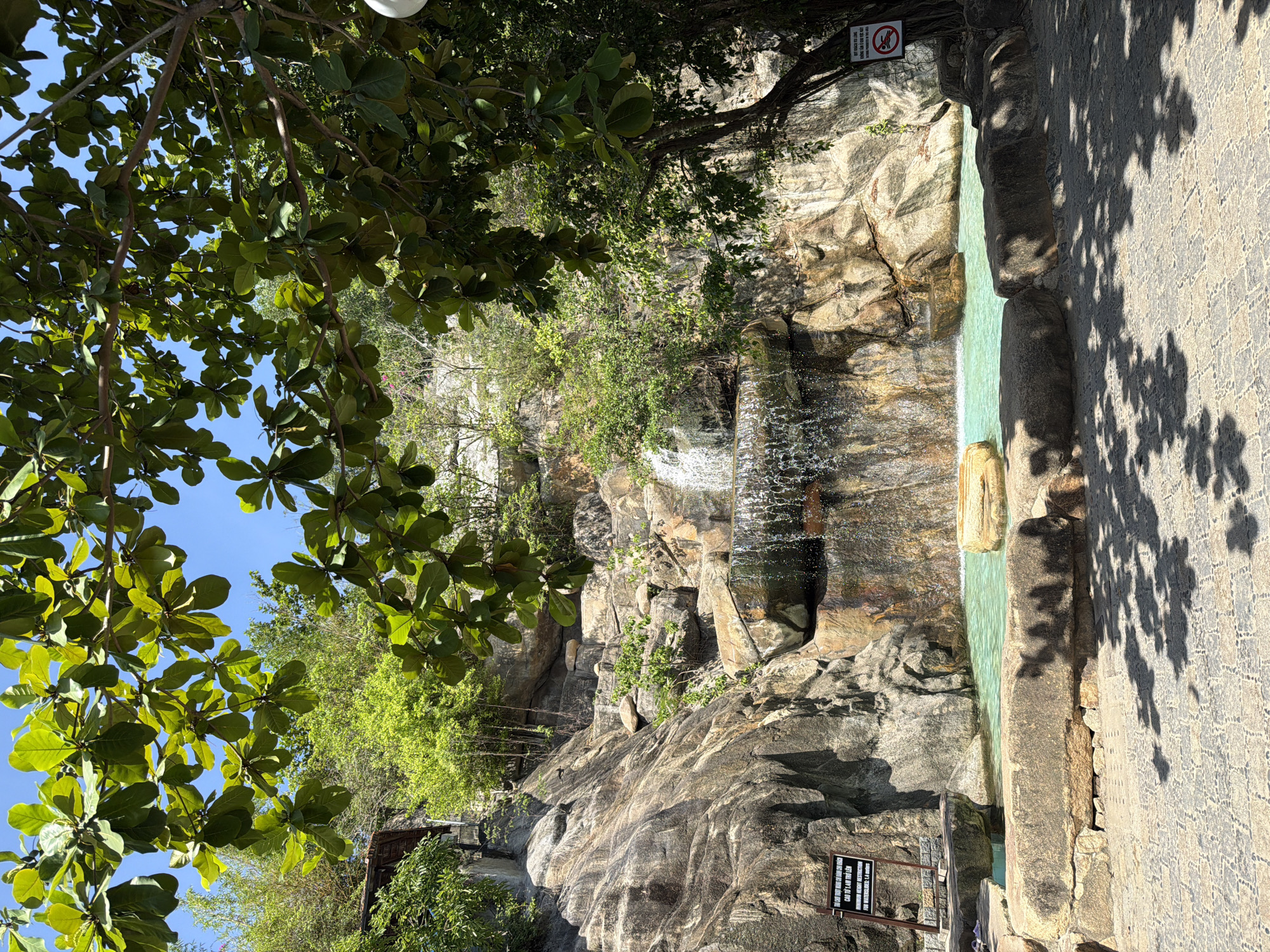 The image depicts a scenic view of a man-made waterfall and pool area, likely part of a resort or park. The waterfall cascades down a rocky incline into a turquoise pool. Lush green foliage and trees frame the scene, with sunlight filtering through the leaves, casting dappled shadows on the paved ground in the foreground. Two signs are visible: one on the left with Vietnamese text and "MINIMUM HEIGHT RESTRICTION FOR WATER SLIDES: 1.4 METERS", and another on the right with a "no swimming" symbol. The setting appears to be outdoors, under a clear blue sky, suggesting a warm climate. The overall impression is one of a tranquil, naturalistic recreational space.