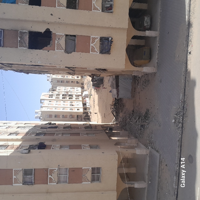 A street view shows damaged buildings on both sides. The structures have multiple floors and are painted in tan. Some windows are missing or broken, and debris litters the ground between the buildings. A distant structure with many floors can be seen at the end of the street. The image is taken from an elevated perspective.
