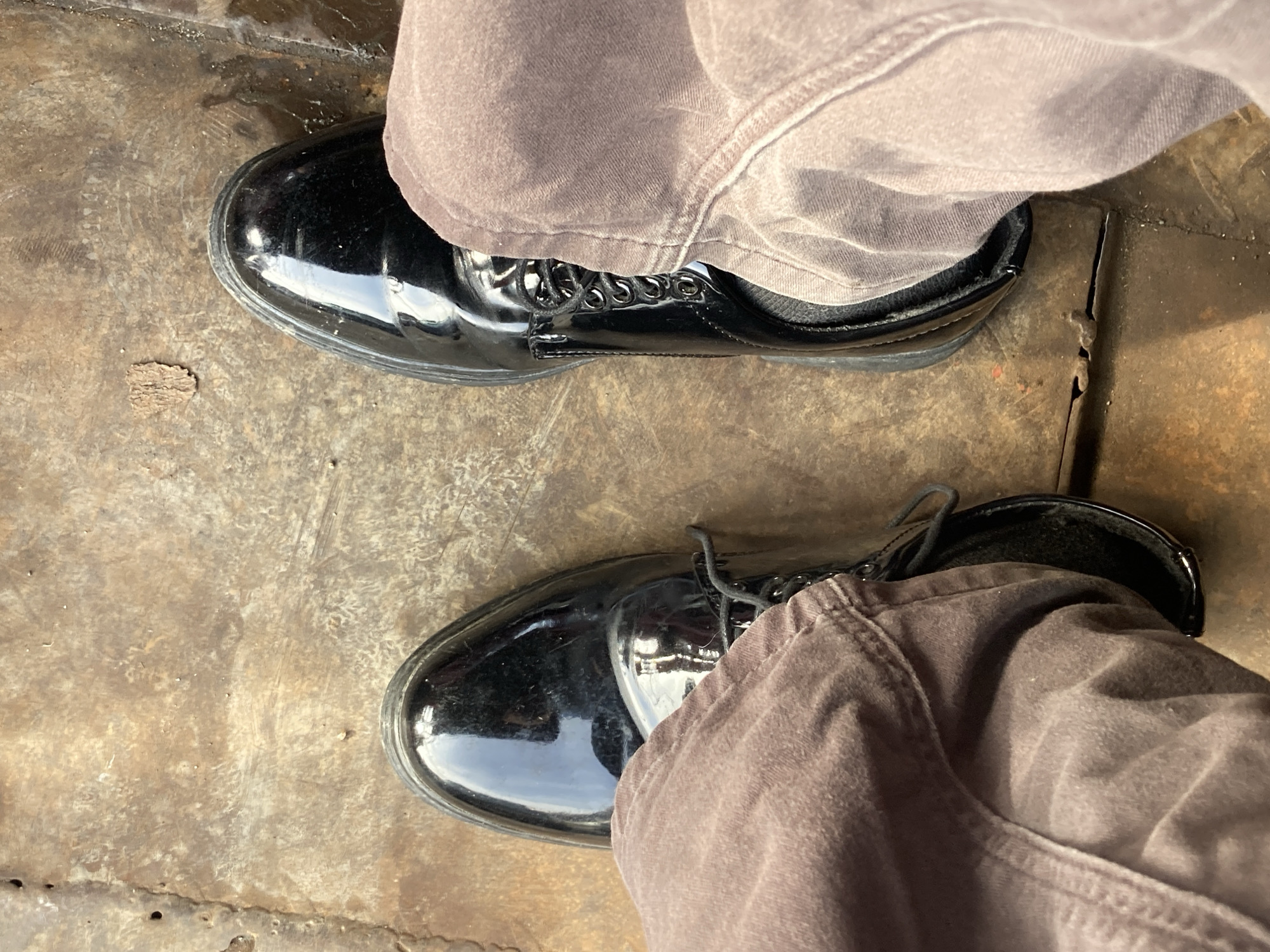 The image is a close-up, top-down view of a person's feet wearing black, shiny, lace-up shoes and loose, light brown or taupe pants. The shoes appear to be dress shoes or work boots. The person is standing on a textured, weathered, and slightly discolored surface, which appears to be a metal grate or flooring. There is some debris on the surface, including a small, dry leaf-like object. The lighting suggests it is daytime, possibly outdoors. The scene does not contain any specific landmarks or indicators to pinpoint its location within Davao City, Philippines.