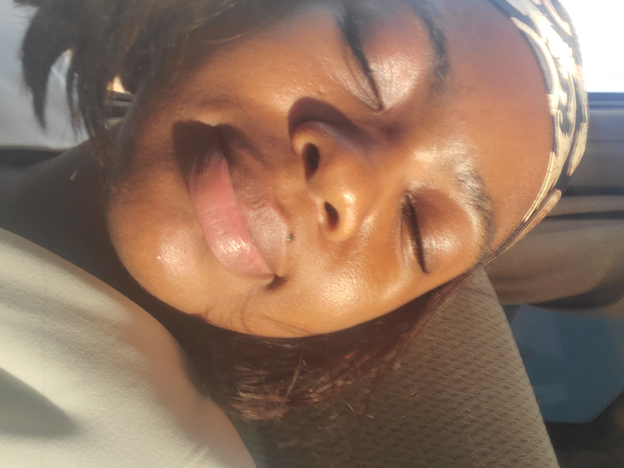A medium-close shot depicts a woman with brown skin lying with her eyes closed. The light reveals that the skin on her face is shiny. She wears a patterned headband. A stud is visible on her lower lip. Her head rests on a dark grey surface, likely fabric, and she is wearing a white shirt. Based on the provided information, it is impossible to verify the location in the city of Kakau, Nigeria.