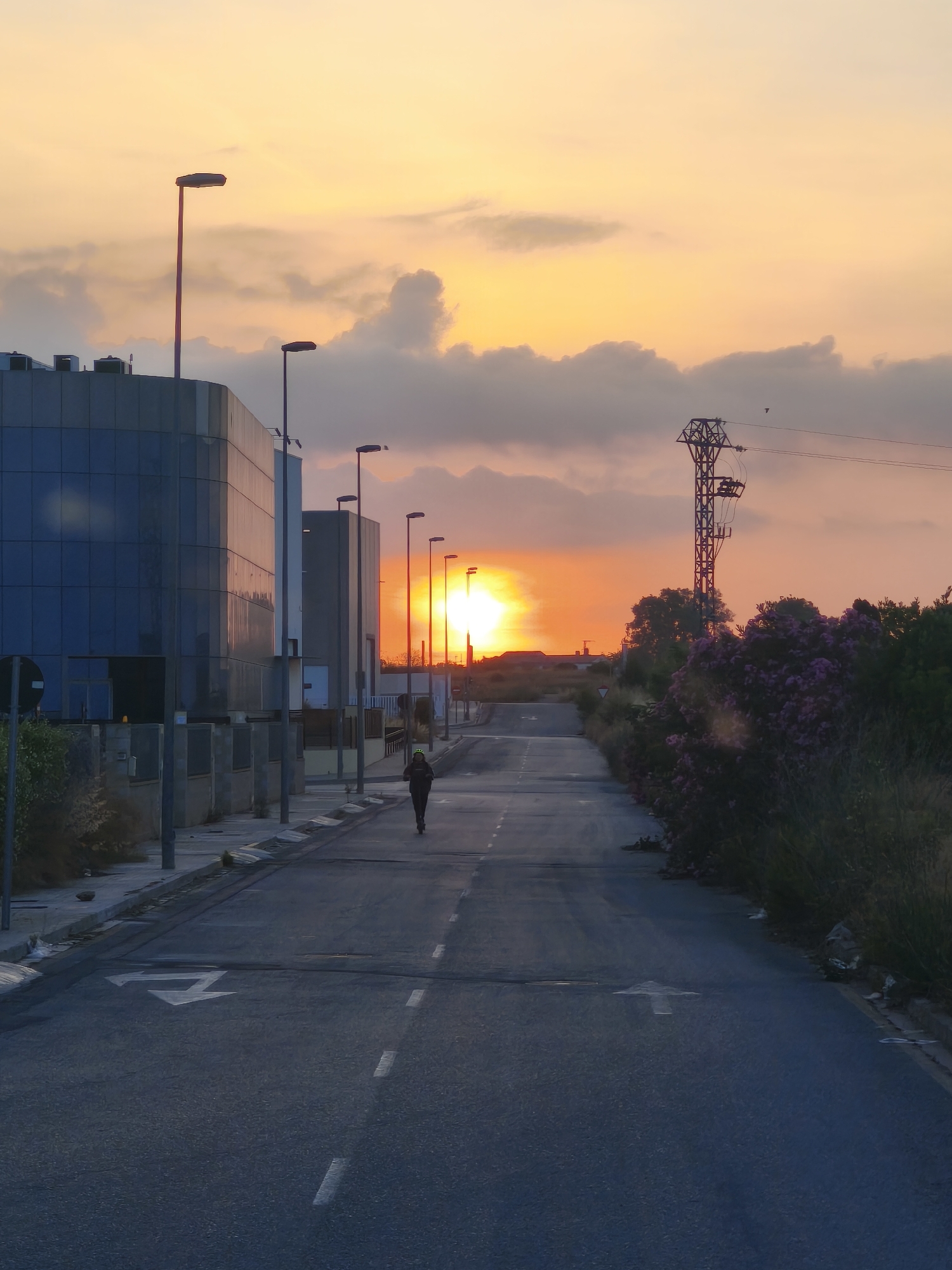 The image depicts a sunset scene in Nàquera, Spain, on a paved road with markings. On the left side of the road, a modern building with blue glass facade stands, bordered by a sidewalk with streetlights. A lone individual, viewed from behind, walks down the center of the road, away from the viewer. The road stretches into the distance, where the sun is setting, casting an orange glow across the sky and illuminating the clouds. To the right of the road, lush green vegetation and flowering plants are visible, along with a tall electricity pylon. The overall atmosphere is serene, with the warm colors of the sunset contrasting with the urban elements.