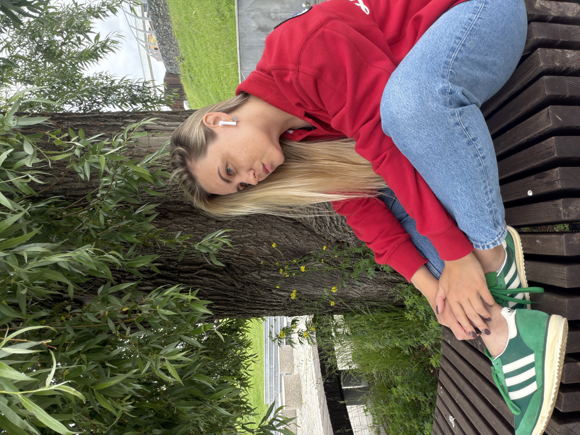 A young woman with blonde hair and blue eyes is sitting on a wooden bench in a park. She is wearing a red jacket and blue jeans. She has green Adidas sneakers on her feet and white earbuds in her ears. The woman is looking away from the camera, to her right. She is sitting under a large tree with green leaves. In the background, there is a grassy hill and a modern building with a curved roof. The scene appears to be located in the city of Votkinsk, Russia.