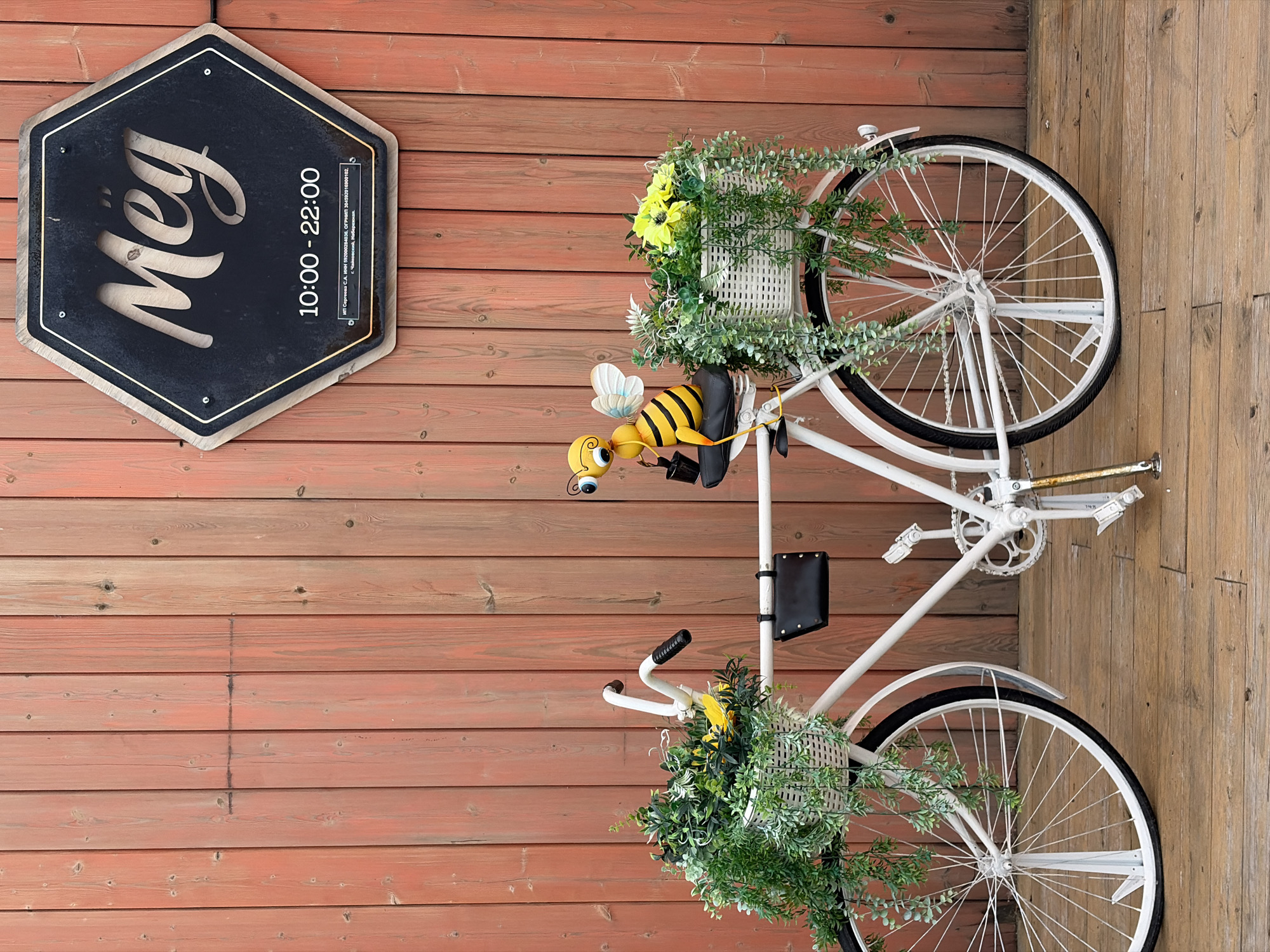 A decorative white bicycle with two wicker baskets filled with artificial plants and flowers is positioned in front of a weathered, reddish-brown wooden plank wall. A cartoonish bee figurine is seated on the bicycle's seat, holding a small black bucket. Above the bicycle, a hexagonal black sign with a gold trim displays the word "Meg" in a stylized font, followed by the operating hours "10:00 - 22:00" and small text including company registration information and "г. Чайковский, Набережная." The ground is a wooden deck. The scene appears to be an outdoor setting, likely an entrance or decorative display for a business in Chaikovsky, Russia.