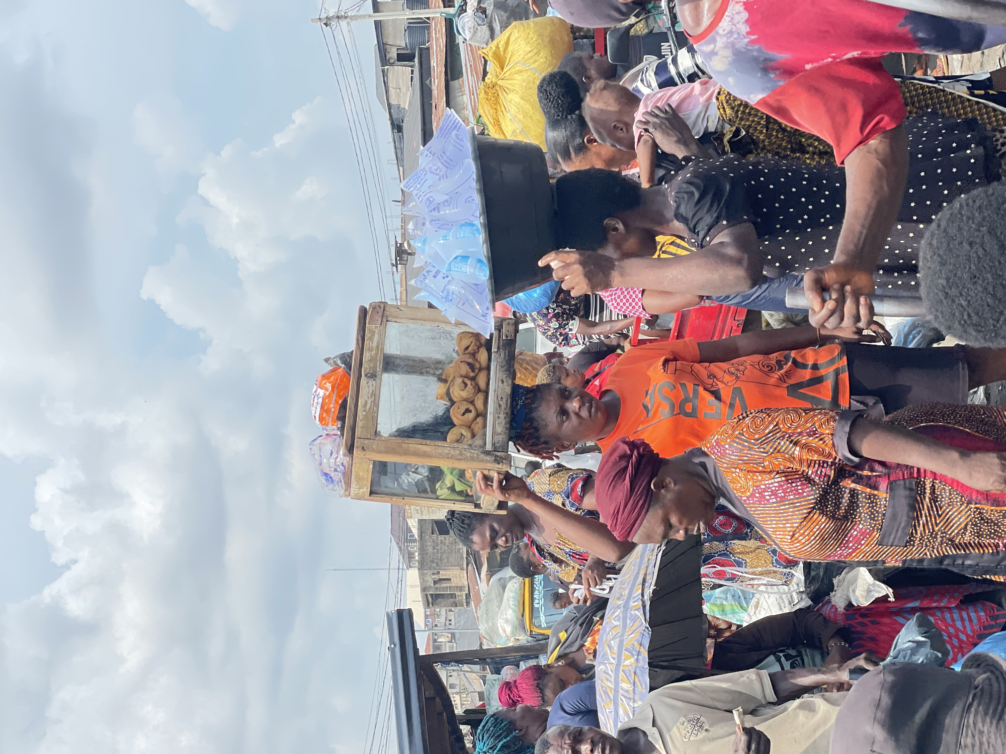 This image captures a vibrant and densely crowded outdoor street scene, likely a bustling market or urban area. The photo is oriented vertically (rotated 90 degrees clockwise), with a bright sky on the left and the busy ground level on the right.
Numerous individuals, primarily adults of African descent, fill the frame. They are closely packed, suggesting high foot traffic. Many are dressed in colorful, patterned fabrics and headwraps, alongside casual t-shirts. Several people appear to be vendors; one individual in the mid-ground balances a large black basin overflowing with clear sachets of water on their head. Another person stands behind a wooden display case with glass panels, which holds numerous small, round, light-brown fried snacks, possibly a local delicacy. Other people are seen navigating the crowd, some holding hands, while others seem to be engaged in transactions or observing the goods. In the foreground (bottom right of the rotated image), a person wears an orange t-shirt.
The setting is an informal urban environment. In the background, indistinct buildings with corrugated metal roofs are visible, along with parts of what appears to be a yellow commercial tricycle (keke). Overhead utility lines stretch across the upper left portion of the image. The time of day is bright daylight, indicated by the clear sky with scattered white clouds and strong illumination throughout the scene. The weather appears fair.
Visible text includes "RSA" (possibly part of "VERSA") on the orange t-shirt. The sachets of water clearly display "PURE WATER" repeatedly, along with other smaller, unreadable brand details. Faint text that might read "EVAN GAL" is visible on the side of the yellow vehicle in the background.