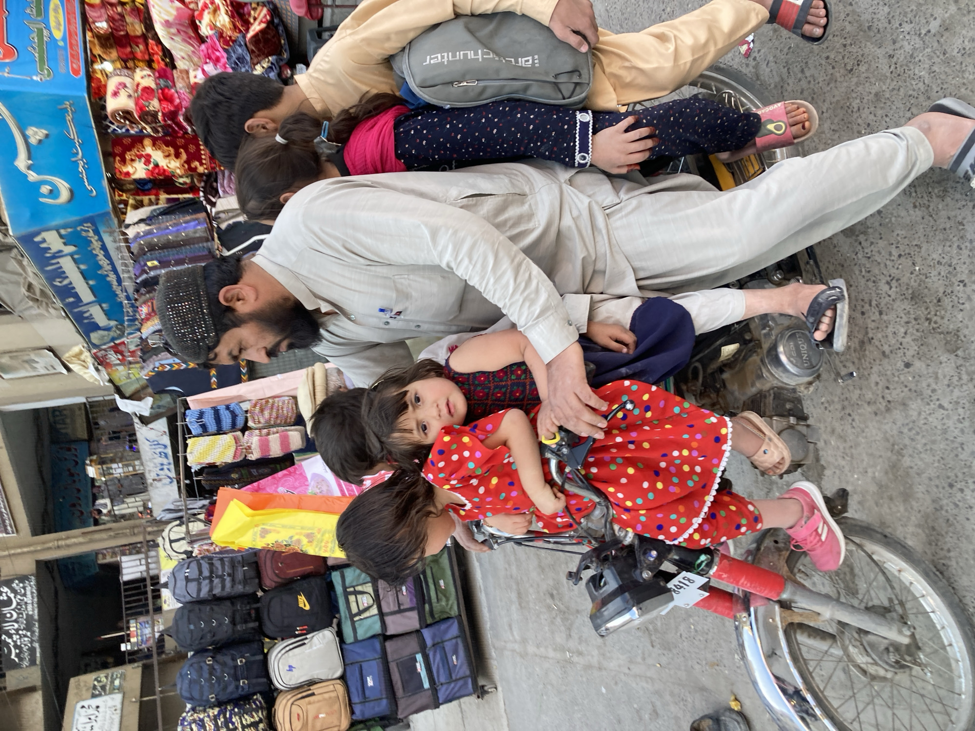 A family, consisting of a father and three children, is crowded onto a motorbike parked on a street in Quetta City Tehsil, Pakistan. The father, with a dark beard and wearing a light tan tunic, is situated on the seat. One child, in a red dress with colorful polka dots, is positioned in front of the father, while another child, wearing a navy top and leggings with a pink scarf, sits behind. A third child, dressed in tan, is at the very back. They are all tightly packed.
The motorbike is old and slightly rusty, bearing the license plate "8418" and the brand "UNIQUE." It is parked on a paved street, likely a market area, as evidenced by the shops and goods displayed in the background.
Various items are visible, including bags, blankets, and other merchandise stacked in front of the shops. A blue sign with white Arabic script is also visible, along with other signs and shopfronts. The overall atmosphere is crowded and busy, typical of a market scene.