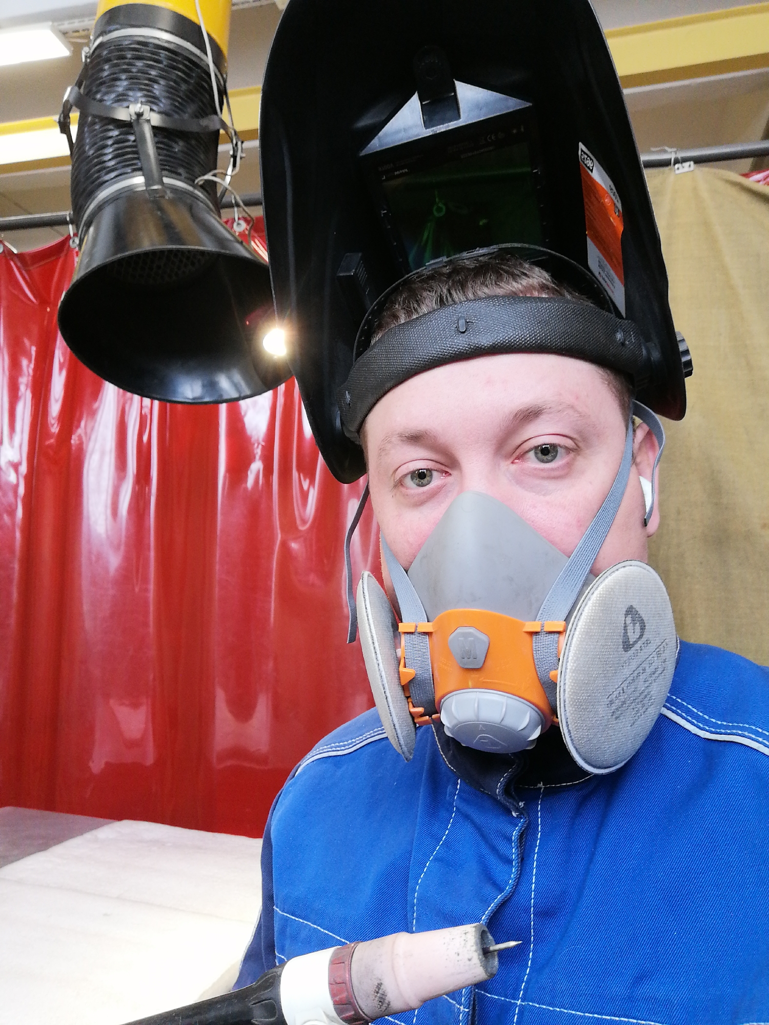 A man is captured in a close-up, eye-level portrait within a welding workshop environment. He has fair skin, short brown hair, and looks directly at the camera with a neutral expression. He is equipped with personal protective gear, indicating he is engaged in or preparing for welding work.
His black auto-darkening welding helmet is flipped up, resting on his head, revealing a dark gray headband. A gray and orange respirator mask covers his nose and mouth, featuring two circular gray filter cartridges on the sides. A white earbud is visible in his right ear. He wears a blue work coverall or jacket with white stitching. In the lower right of the frame, he holds a TIG welding torch, characterized by its white ceramic nozzle and a visible tungsten electrode.
The background shows elements typical of a fabrication or training facility. A black, funnel-shaped fume extractor hood, attached to a yellow flexible duct, is prominent on the left. A bright light source emanates from near the extractor's opening. Behind the man, a vibrant red, translucent plastic or vinyl curtain is visible, commonly used to delineate welding booths. Overhead, fluorescent lighting fixtures suggest an indoor setting. Visible text includes "esab" (partially seen) on the welding helmet and an "M" on the respirator's central component, with additional faint text on the filter cartridges. The scene depicts a welder paused in his workspace.