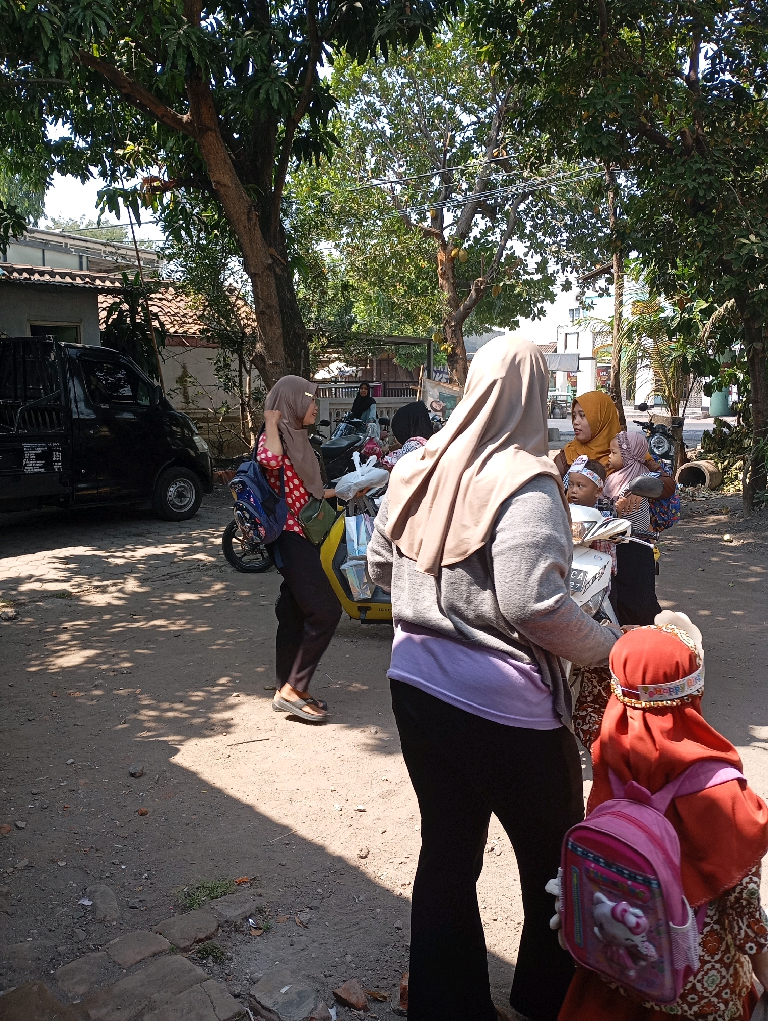 A medium shot captures a sunny outdoor scene with several people, likely in Ketegan, Indonesia, near a dusty dirt road with scattered debris. In the foreground, a child wearing a red hijab and a pink Hello Kitty backpack stands with their back to the camera. To their left, a woman in a light brown hijab and grey sweater, with a purple undergarment, faces away from the viewer, holding onto a white scooter. Behind her, a woman in a red and white polka dot shirt and a tan hijab, carrying a blue backpack, walks away from the scooter. Further back, a woman in a yellow hijab holds a baby, and another child sits on the scooter. A black pickup truck is parked to the left of the frame. Trees with green foliage provide shade, casting dappled shadows on the ground. The background shows a house with a tiled roof and power lines.