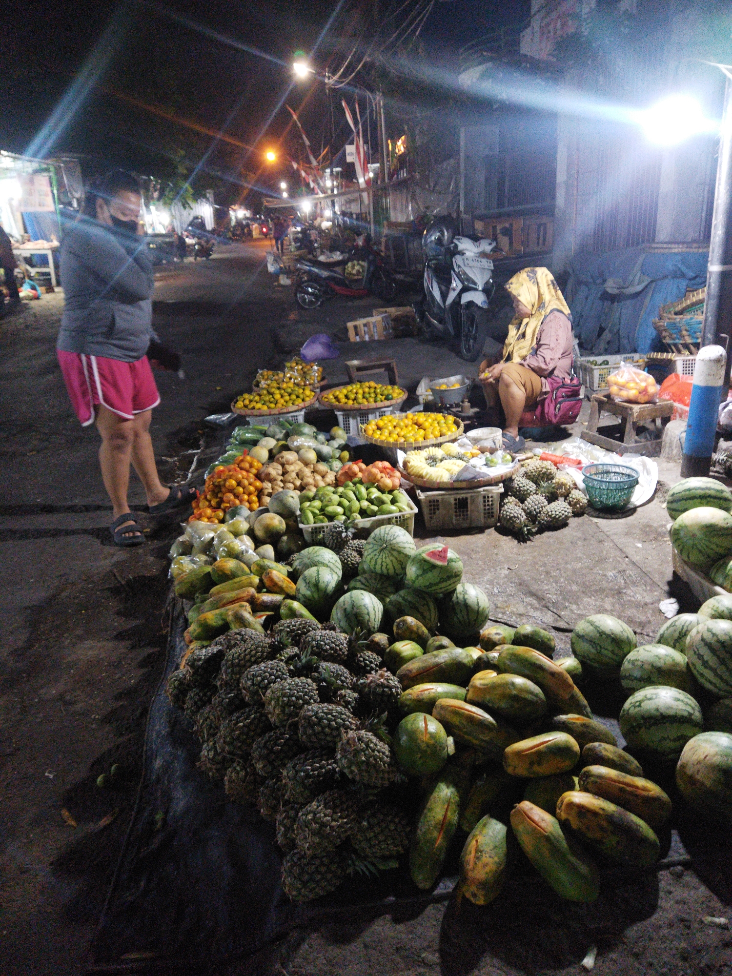 A nighttime street market scene in Semarang, Indonesia, is depicted. A person wearing a grey hoodie and pink shorts stands on the left, facing towards the right, with their arms crossed and a black face mask on. To the right, a woman in a yellow headscarf and a pink shirt sits near a stall. The market displays a variety of fruits, including pineapples, watermelons, papayas, and small oranges, arranged on tables and the ground. Several motorcycles are parked in the background, and streetlights cast a bright glow on the scene. The ground is wet, suggesting recent rain.