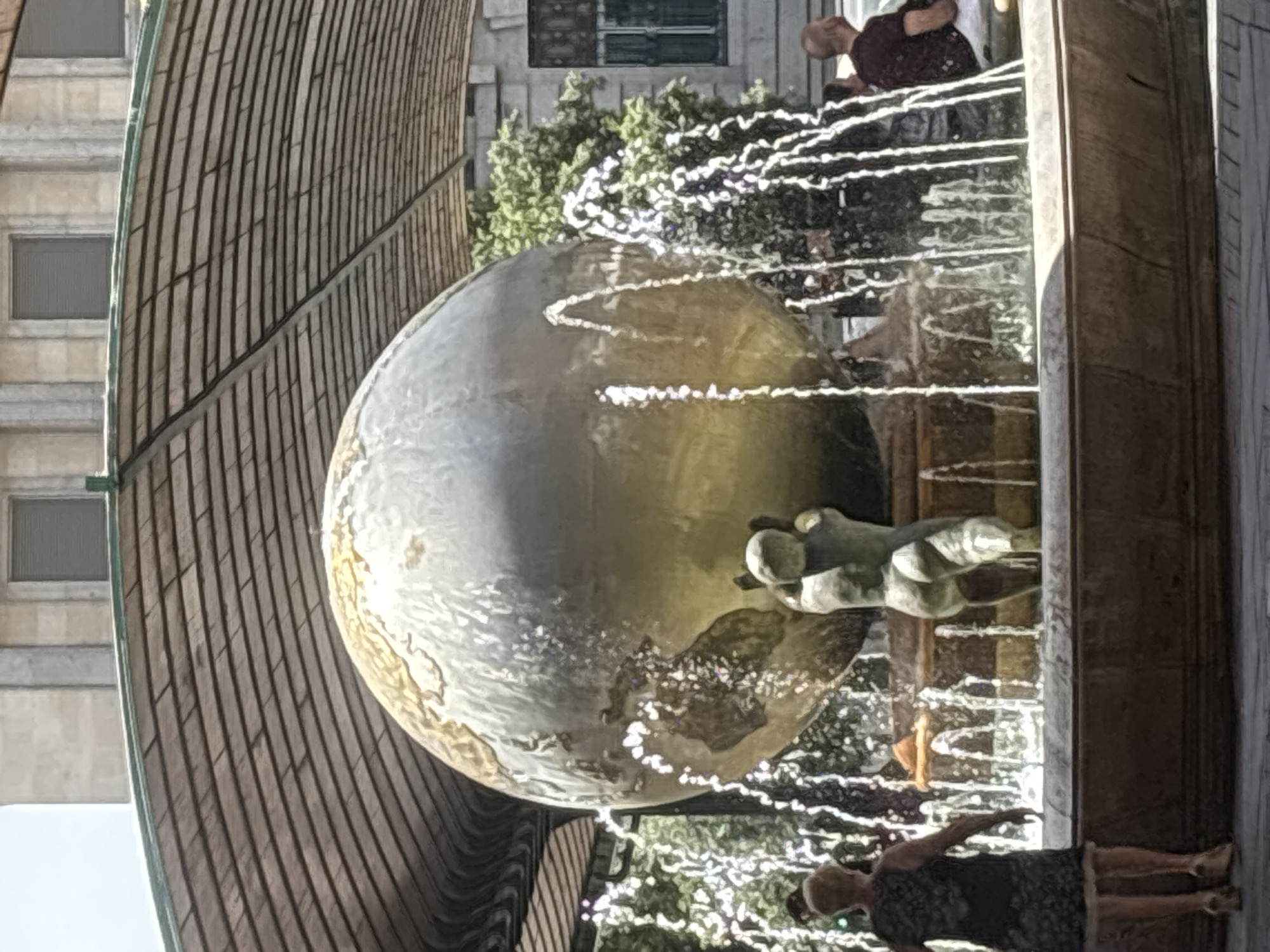 In Valladolid, Spain, a large globe sculpture, possibly representing Earth, is a central feature of a public fountain. The globe is positioned on a pedestal and appears to be supported by a smaller sculpted figure underneath. Water jets erupt around the globe, creating a dynamic visual effect. The fountain is situated outdoors, beneath a distinctive arched canopy constructed with rows of dark, rectangular elements. In the background, a stone building with windows is visible, along with lush green trees. Several people are present, with one woman in the foreground looking away from the camera, and others in the background observing the fountain. The overall scene captures a moment in a public space, likely a plaza or park, with artistic and architectural elements combining to create an engaging environment.