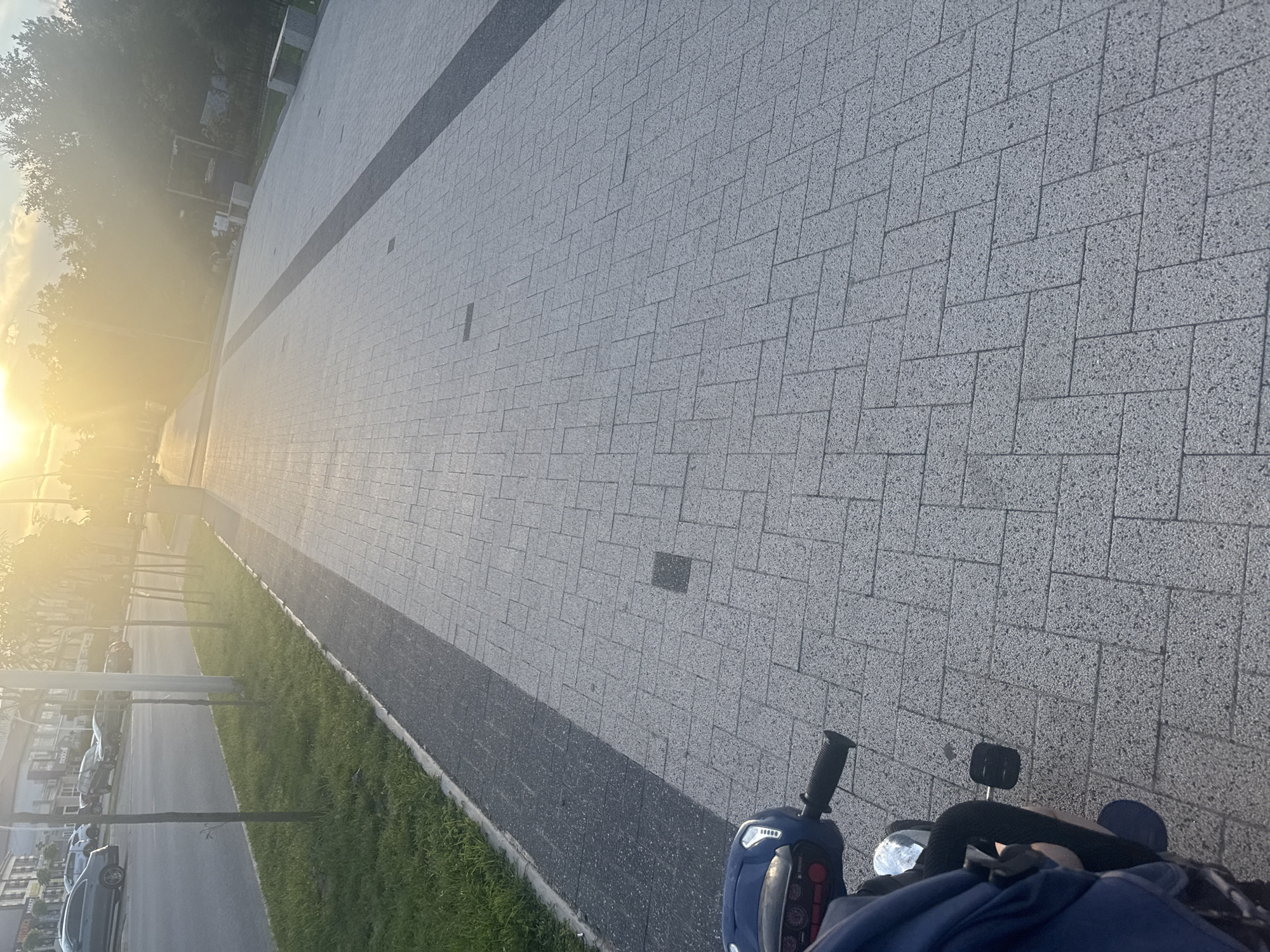 The image shows a view from a scooter moving along a paved pedestrian walkway. The walkway is made of gray interlocking rectangular pavers arranged in a herringbone pattern. A darker gray strip runs down the center of the walkway. To the left of the walkway is a strip of green grass, and beyond that is a street with several parked cars. In the background, there are buildings and trees, and the sun is setting, casting a bright, hazy light across the scene. The scooter is blue and has a handlebar with controls and a pedal visible. A person's legs, dressed in dark clothing, are visible on the scooter. The location is an urban outdoor environment during sunset.