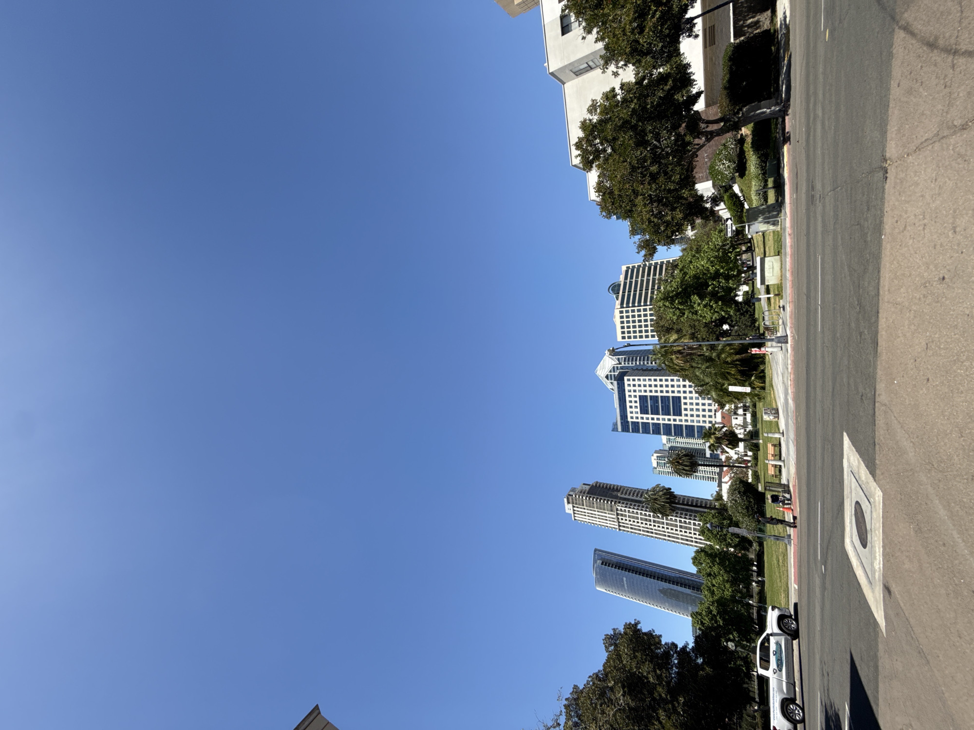 The photograph shows a street scene in San Diego, California, United States. The view is oriented upwards towards the sky, revealing several tall buildings lining a street with sidewalks, green spaces, and trees. Cars are parked along the curb. A person is walking on the sidewalk. The sky is a clear blue.