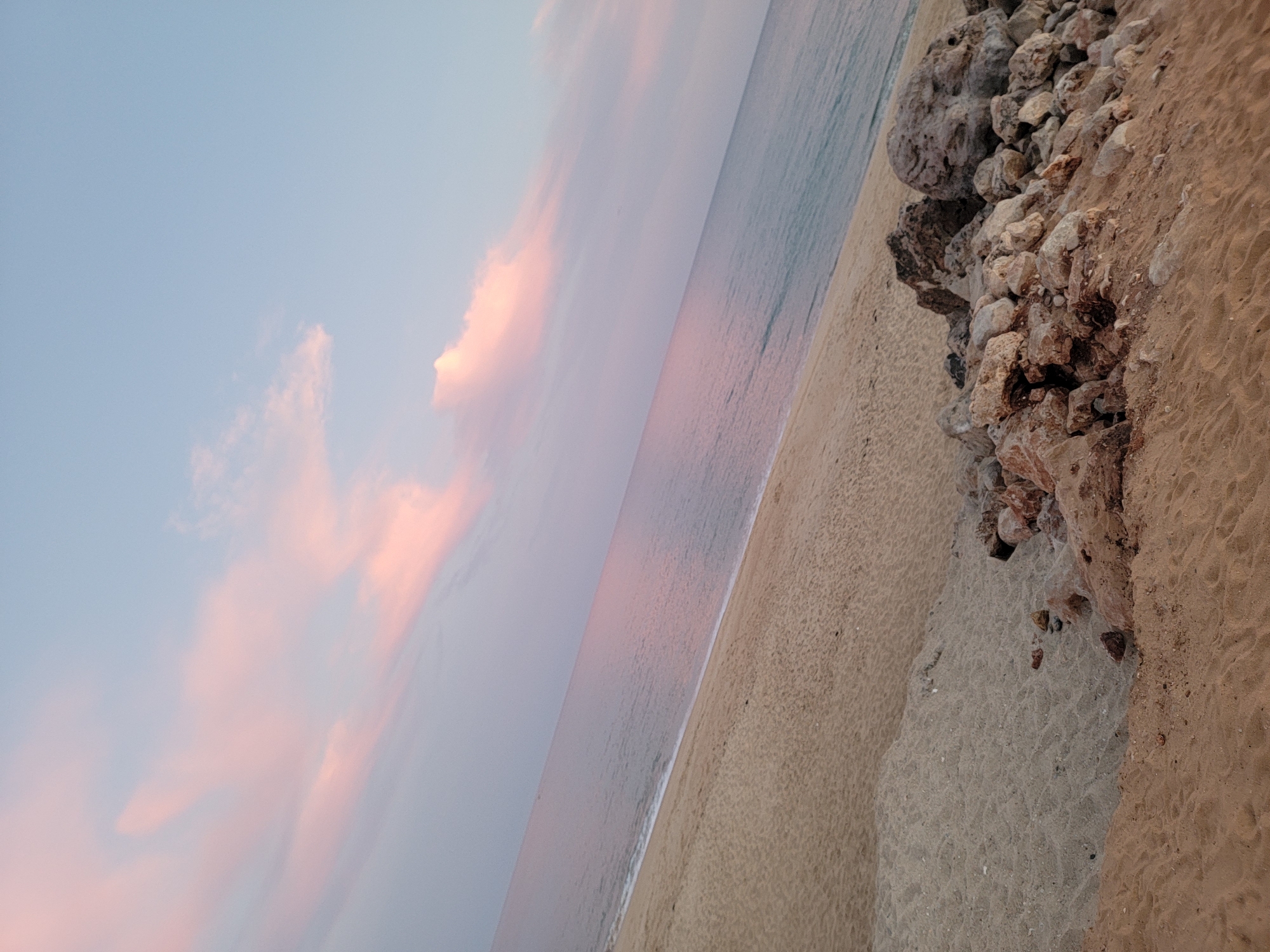 The image shows a beach with sand, rocks, and the ocean. The sky is a soft blue with streaks of pink clouds, suggesting it is either sunrise or sunset. The water is calm and reflects the colors of the sky. There are a pile of rocks in the foreground on the sand. The overall scene is serene and natural. This scene could be located on any of the beaches in Quarteira, Portugal, known for its coastline and beautiful sunsets.