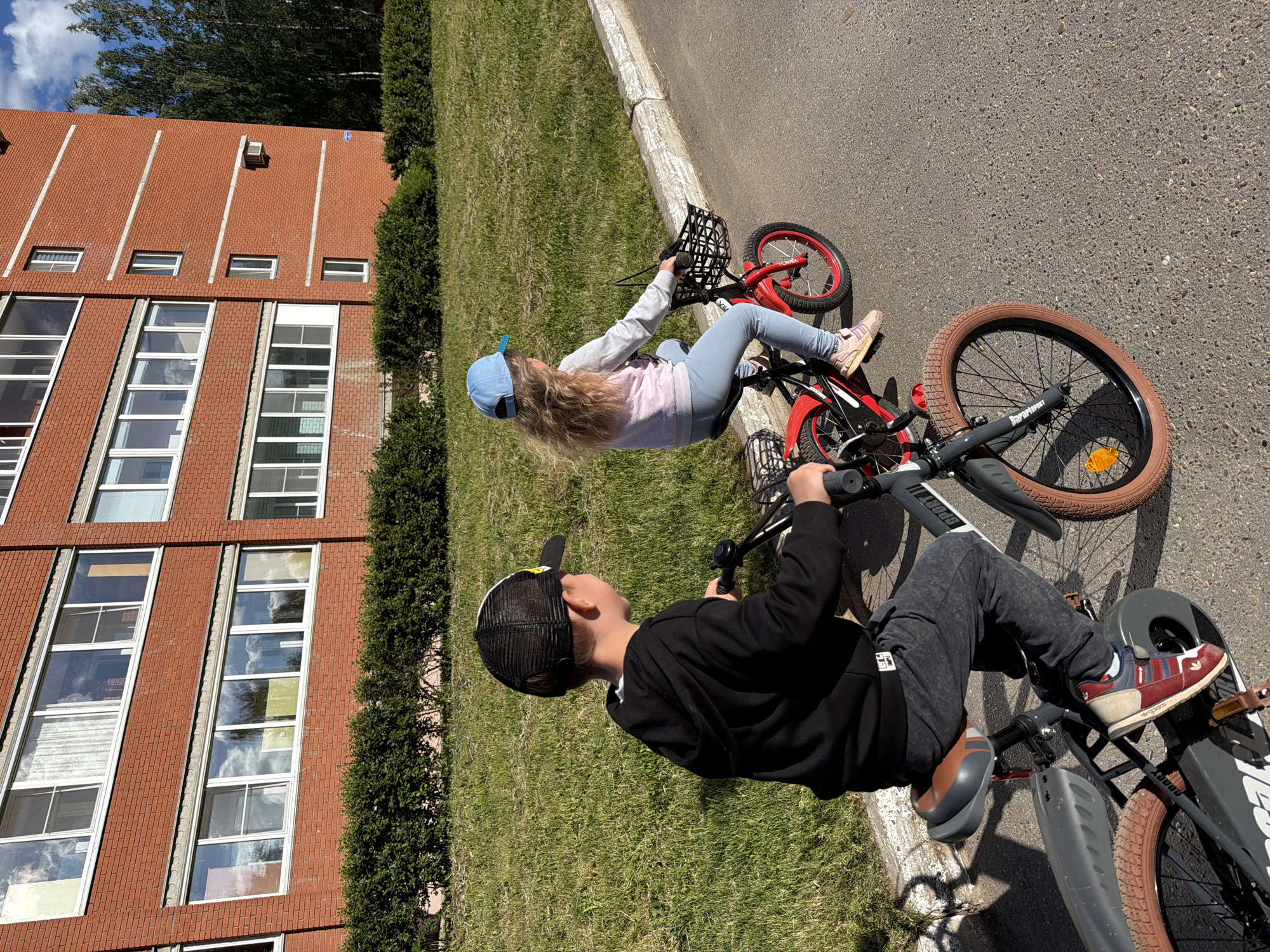 Two children are riding bicycles next to a red brick building in Chaikovsky, Russia. A girl with long blonde hair wearing a blue baseball cap, light pink long-sleeved shirt, and light blue leggings is riding a red bicycle with a black basket attached to the front. A boy is riding a gray bicycle with tan tires and is wearing a black long-sleeved shirt, gray sweatpants, a black baseball cap with white mesh on the back, and multi-colored sneakers. The building behind the children is tall, with a red brick facade and multiple windows. There is a patch of grass between the road and the building, and a line of trees can be seen in the background.
