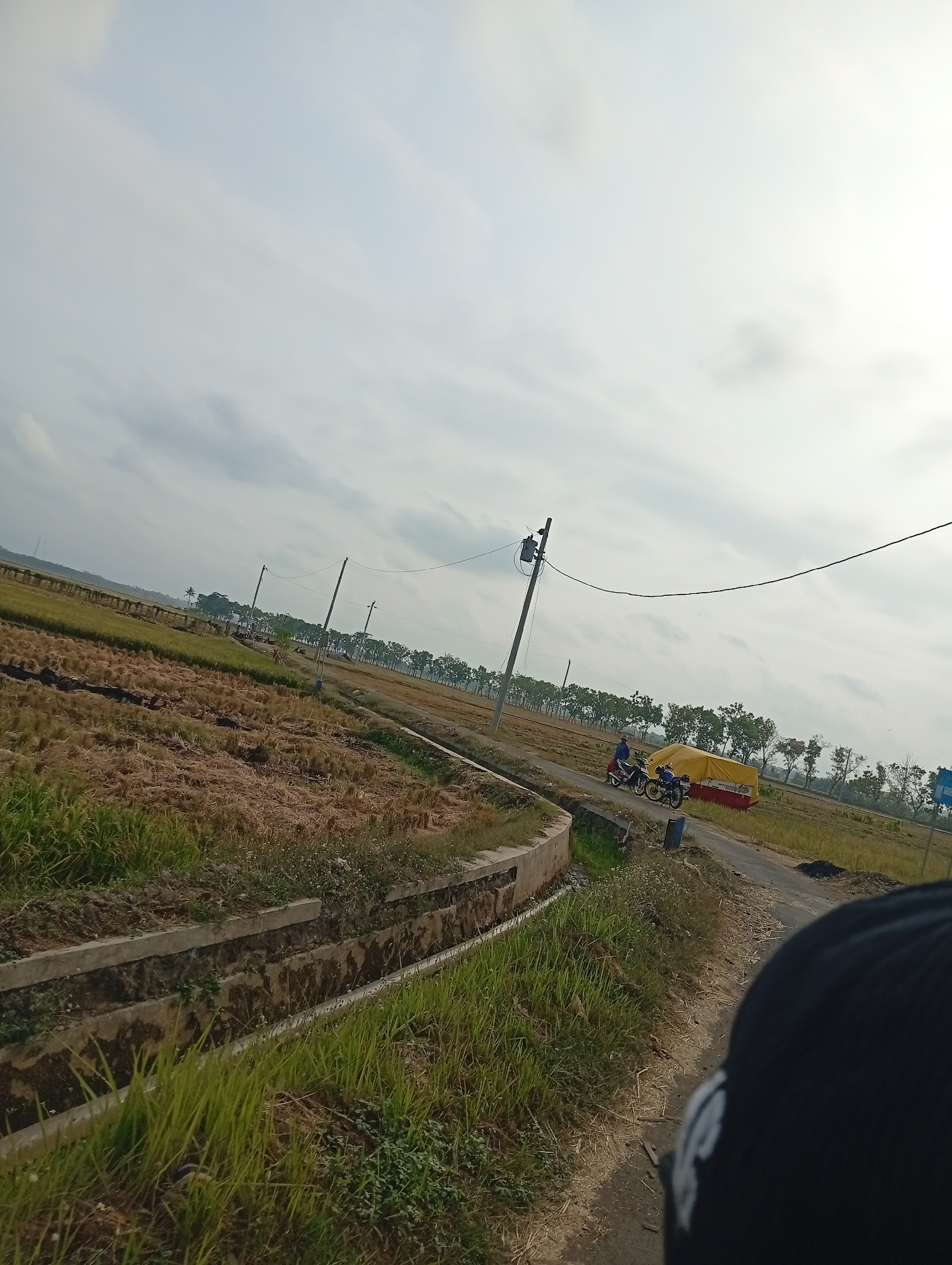 The image depicts an outdoor scene with a vast, overcast sky. In the foreground, a person's head, wearing a dark baseball cap, is visible on the right side. A dirt path runs alongside a grassy embankment, which features a concrete-lined drainage ditch. Beyond the ditch, a harvested field, characterized by dry, stubbly vegetation, stretches into the distance. A road borders the field, and on the road, two motorcycles are parked next to a vehicle covered with a bright yellow tarp. Several utility poles with wires are scattered throughout the landscape, and a line of trees forms a distant horizon. The overall setting appears to be rural.