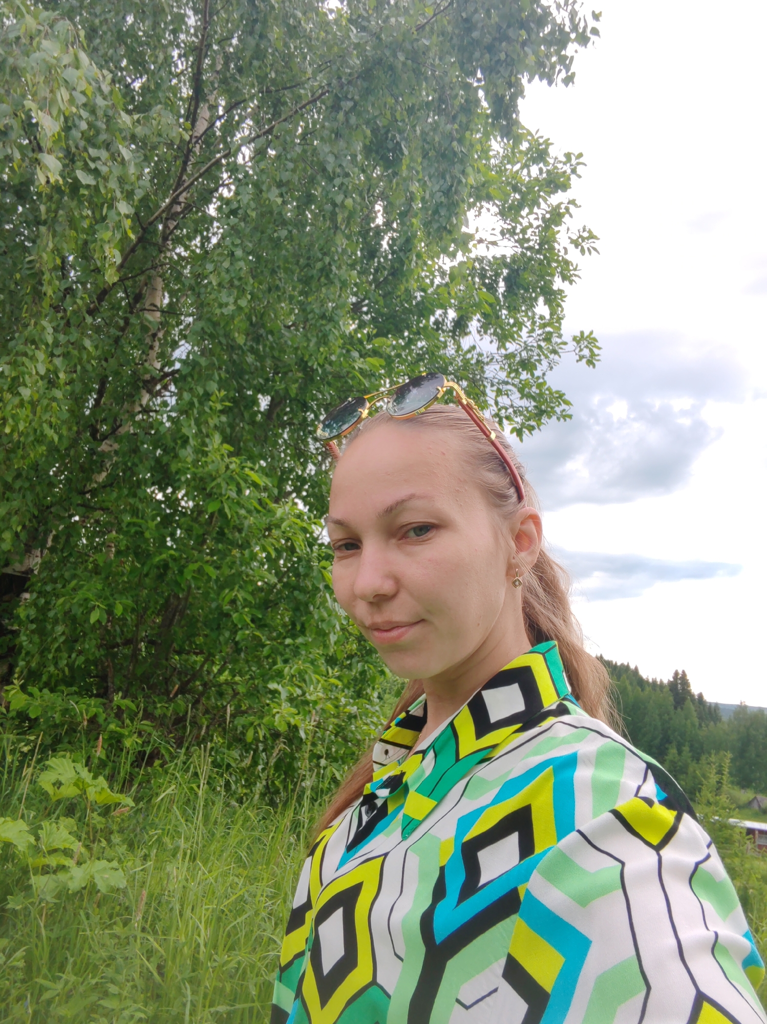 A young Caucasian woman with fair skin and light brown hair pulled back into a ponytail is posing for a selfie outdoors, with a backdrop of lush green trees and a cloudy sky. She is wearing sunglasses on top of her head and a brightly colored, geometric-patterned shirt in shades of green, blue, yellow, and black. The woman is looking directly at the camera with a slight smile. The scene appears to be a natural setting, possibly a park or a rural area, with tall grass and trees surrounding her. The lighting suggests it is daytime, with soft, diffused light. There is no specific indication of the city of Чухлэм, Russia, in the image itself, as the background is generic nature.