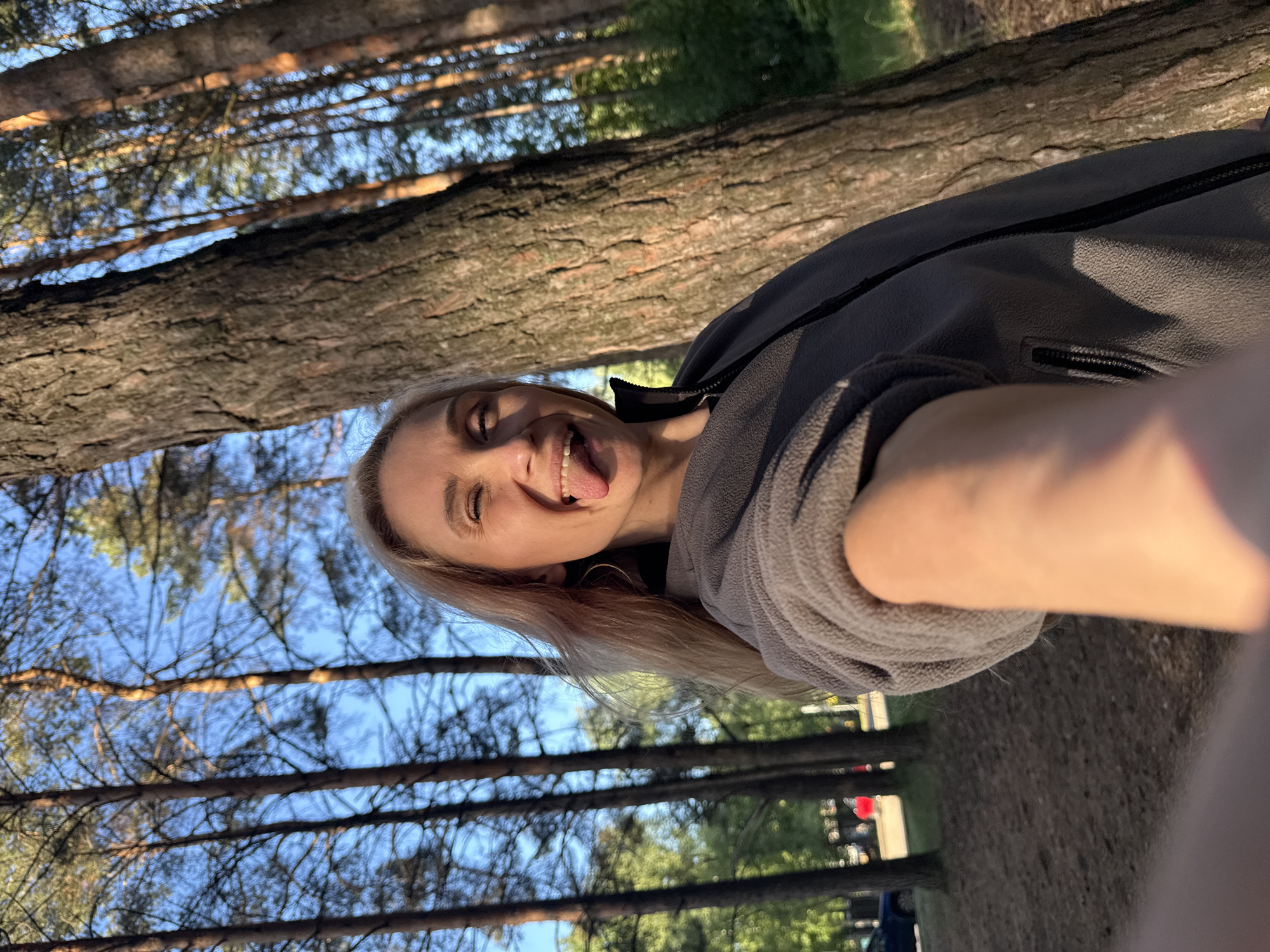 The image captures a selfie of a woman in an outdoor, wooded environment, likely in Chaikovsky, Russia. The photo is rotated 90 degrees counter-clockwise. The woman, with blonde hair and wearing a grey fleece jacket, smiles playfully at the camera, winking her left eye and sticking out her tongue, conveying a lighthearted and happy mood. Her right arm, holding the camera, extends towards the bottom right of the frame.
The background features numerous tall tree trunks with textured brown bark, appearing horizontally across the frame due to the image's orientation, suggesting a forest or park setting. Patches of clear blue sky are visible through the tree branches and foliage. The warm sunlight indicates it is daytime, possibly late afternoon or early morning, casting highlights on the trees and the woman. No visible text is present in the image.