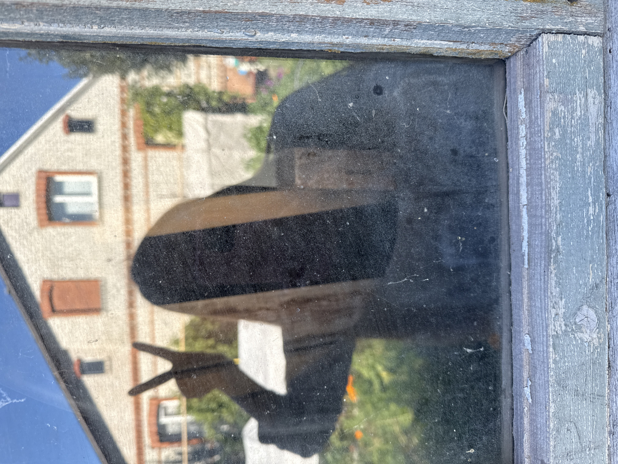 A reflection of a person making a peace sign is visible on a dirty window pane. The person's face is obscured by shadow, but their arm and hand are clear. Behind the person, the reflection shows a light-colored house with orange window frames and a peaked roof, set against a blue sky. The house appears to be in a residential area. The window is set into weathered, light blue wooden framing, suggesting an older structure. The image captures an outdoor scene in Zheleznogorsk, Russia, likely a residential property, as indicated by the house and garden visible in the reflection.