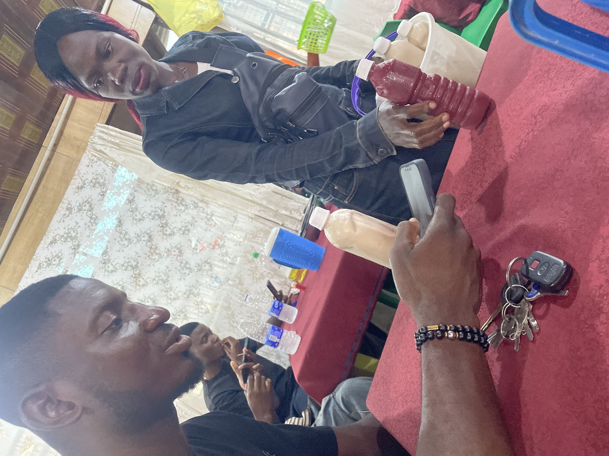 The image depicts an indoor scene, possibly a restaurant or a gathering place, with a focus on individuals and items on a table. In the foreground, a person's arm with a bracelet made of black beads and gold accents is visible, holding a set of keys with a car remote. The hand is resting on a red tablecloth. To the left, the profile of a man's face is visible, looking downwards. Behind him, another man is also looking down and appears to be eating.
On the red table, there are several bottles of water, one blue plastic cup, and two bottles containing beverages. One bottle contains a creamy-colored liquid, and another contains a reddish liquid. A white bucket is also on the table, filled with similar-looking bottles.
In the background, a woman with dark hair and streaks of red is wearing a dark denim jacket over a white top and jeans. She has a dark fanny pack worn across her chest. She is looking towards the right side of the frame. The background also shows a patterned curtain and wooden ceiling beams, suggesting an interior setting. The presence of these elements indicates a casual, social gathering. While the image was provided with the context of Abuja, Nigeria, there are no explicit visual cues within the image itself that definitively confirm this specific location.