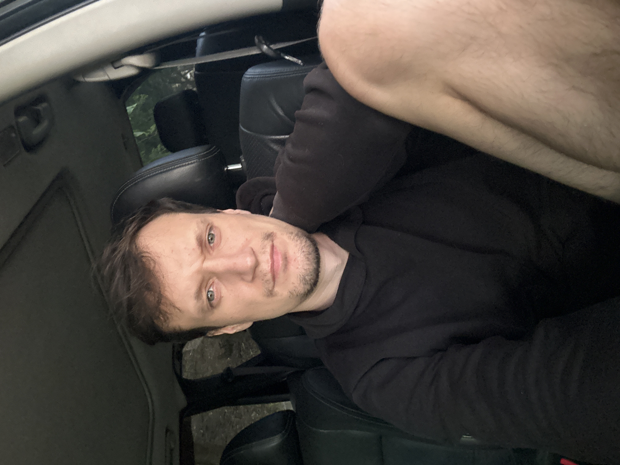 A young Caucasian man is seated inside a car, possibly in Makiv, Ukraine, given the context. He has short, dark brown hair and a light beard. He is wearing a black hooded sweatshirt. His right arm is resting on his lap, and his left arm is bent with his hand near his neck. The interior of the car appears to be black leather. Sunlight filters in from the top, suggesting it is daytime. The background is blurred, but appears to be foliage.