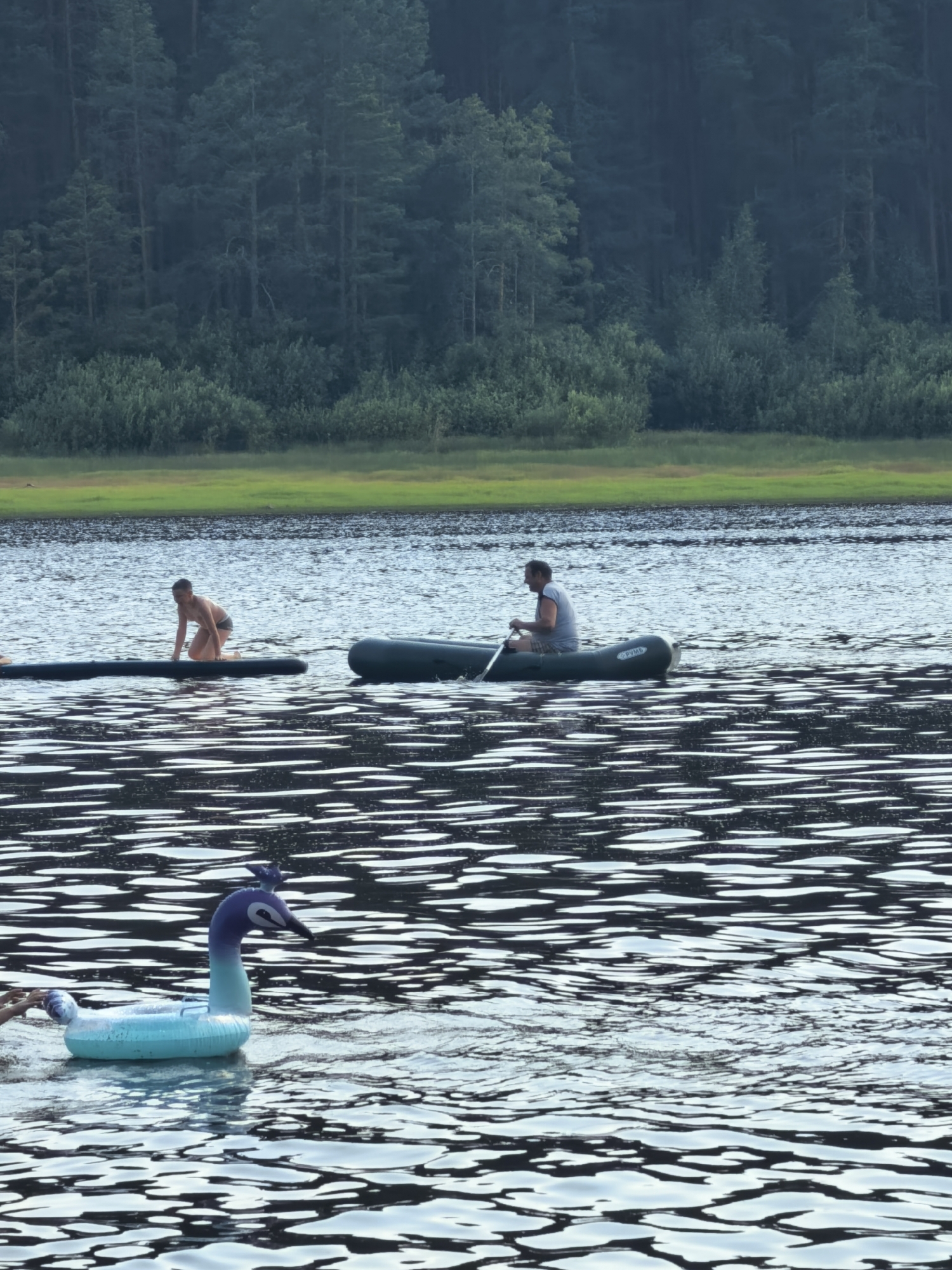 In the city of Sysert, Russia, on a body of water, a man is paddling an inflatable boat with a single oar. In the background, a person kneels on an inflatable raft. Closer to the foreground, an inflatable toy shaped like a peacock floats on the water. The scene is framed by a dense forest on the distant shore.