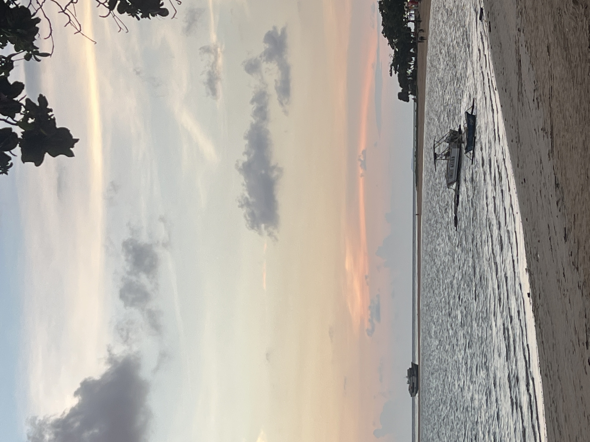 The image depicts a serene beach scene at dusk in Cuyo, Philippines. The sky is painted with soft hues of pink, orange, and pale blue, with scattered clouds. In the distance, a large cargo ship is visible on the horizon. Closer to the shore, two smaller boats are anchored in the calm water, one with "LIFE GUARD" written on its side. The beach is sandy, with the gentle waves of the sea lapping at the shore. The silhouette of trees and a few people walking can be seen along the coastline in the background, adding a sense of quiet activity to the peaceful atmosphere.