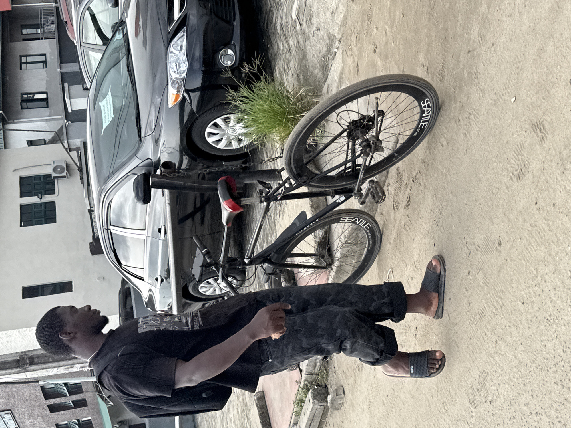 An outdoor scene in Ado, Nigeria, shows a man standing next to a bicycle with parked cars and buildings in the background. The man, positioned on the right, is seen in profile, facing left. He has dark skin, short dark hair, and a short beard, dressed in a dark short-sleeved collared shirt, dark camouflage-patterned pants, and black slides. His right hand is near his hip.
A black bicycle stands upright to his left, featuring "SEATTLE STYLE" written in white on its black rims and a red and black seat. Behind the bicycle and man, a black car (possibly a Toyota Corolla) is parked. A lighter-colored car is parked behind the black one, displaying a white "FOR SALE" sign on its windshield. Several multi-story buildings, mostly light-colored with dark windows, form the background, one showing an air conditioning unit.
The ground is unpaved, appearing sandy or dusty, with visible tire tracks and footprints. A small clump of green grass grows from the ground between the black car and the bicycle. The lighting suggests it is daytime, possibly an overcast day, given the soft illumination and lack of harsh shadows. The man appears to be standing still, with a neutral expression, possibly resting or waiting.