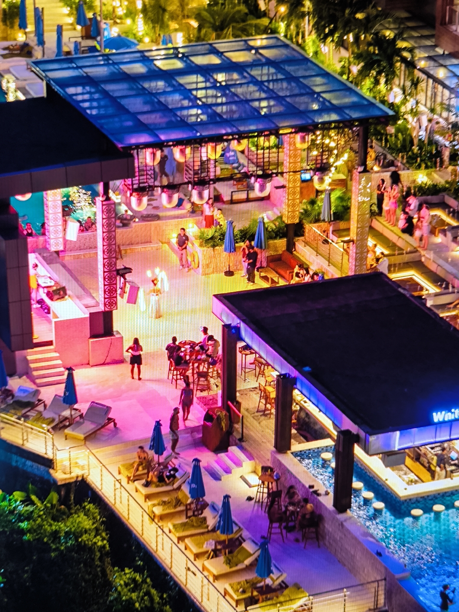 This image depicts a multi-level outdoor entertainment complex at night in the city of Unknown, Unknown. The scene is illuminated by diverse artificial lighting, primarily vibrant pink, purple, blue, and warm yellow. In the upper-left, a large structure features a translucent blue, possibly glass, roof and an open-air area below with ornate pillars and suspended lighting. Within this area, several individuals are visible, some on an elevated platform suggesting a performance or DJ booth.
The central area is an expansive pink-lit patio where approximately twenty people are standing, walking, or seated at tables and high stools. One individual in white attire stands centrally, seemingly performing or posing. Another person, wearing a dark top and light shorts, stands with hands on hips.
To the bottom right, a dark-roofed structure houses a swim-up bar. The bar features a pool with blue mosaic tiling and several white submerged stools, where patrons are visible. The exterior of this bar structure is edged with blue and purple LED strips, with the word "Wai" partially visible.
Along the lower left and right edges, multiple terraced levels are furnished with numerous light-colored sun loungers, each accompanied by a folded blue umbrella. Several individuals are relaxing on these loungers. Green foliage is visible along the periphery of the complex. Railings separate the various levels and pathways. The overall atmosphere suggests a lively, social gathering or event at a resort or nightclub.