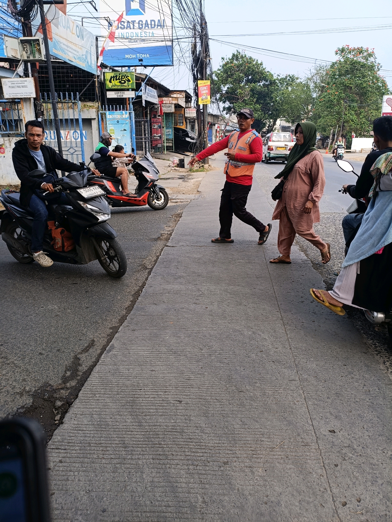 The image captures a street scene in Tangerang, Indonesia, with a focus on pedestrian and vehicle activity on a sunny day. In the foreground, a man is seated on a black motorcycle, looking towards the camera. Another person is visible riding a red and black motorcycle behind him.
Further up the street, a man in an orange vest and dark pants is directing traffic, with his left arm extended. A woman wearing a green headscarf and a light pink dress is walking past him, looking slightly away. Another woman, partially visible in the lower right of the frame, is also on a motorcycle, wearing a light blue scarf and white pants.
The environment includes a concrete sidewalk with textured lines, a paved road, and various buildings and signage in the background. Prominent signs include "PUSAT GADA INDONESIA" and "MOH TOHA," suggesting commercial establishments. Trees with red flowers are also present. Overhead, a complex network of electrical wires crisscrosses the sky. The overall impression is a bustling urban street with everyday activities unfolding.
