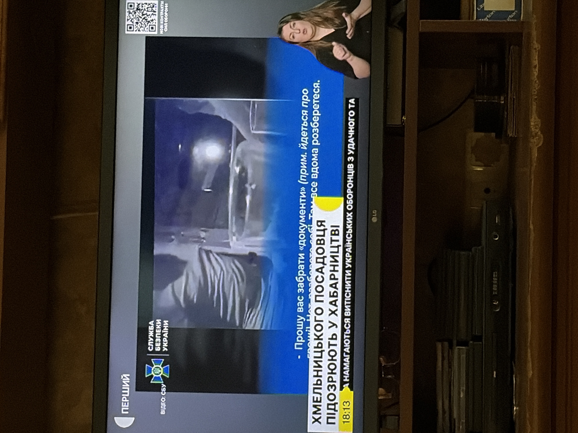 The image shows a television screen displaying a news broadcast. The broadcast includes a video feed, text overlays, and a woman signing in sign language. The text overlays indicate that the broadcast is from "Pervyy" (First) channel and features the logo of the "Security Service of Ukraine." A news ticker at the bottom of the screen reads "Khmelnitsky official suspected of bribery" and "They are trying to drive out Ukrainian defenders from Avdiivka." The screen also shows a timestamp of "18:13". The television is placed on a wooden shelf unit, with a DVD player and stacks of discs visible below it. The background of the room is dark and out of focus.