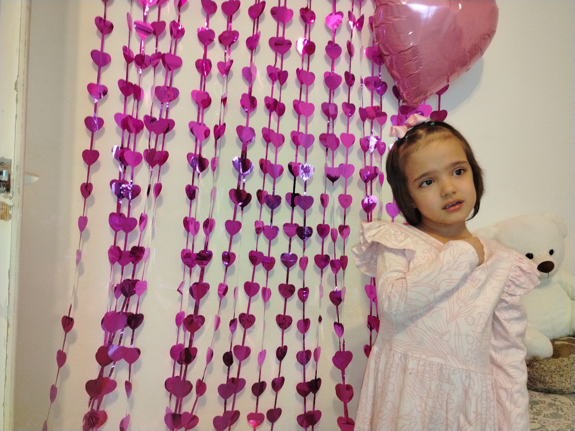 A young girl stands in front of a wall decorated with pink heart-shaped garlands and a pink heart-shaped balloon. The girl is wearing a light pink dress with ruffled sleeves. To her right is a white teddy bear sitting on a textured surface. The location is likely indoors. The city of Noviy, Russia is not evident in the scene.