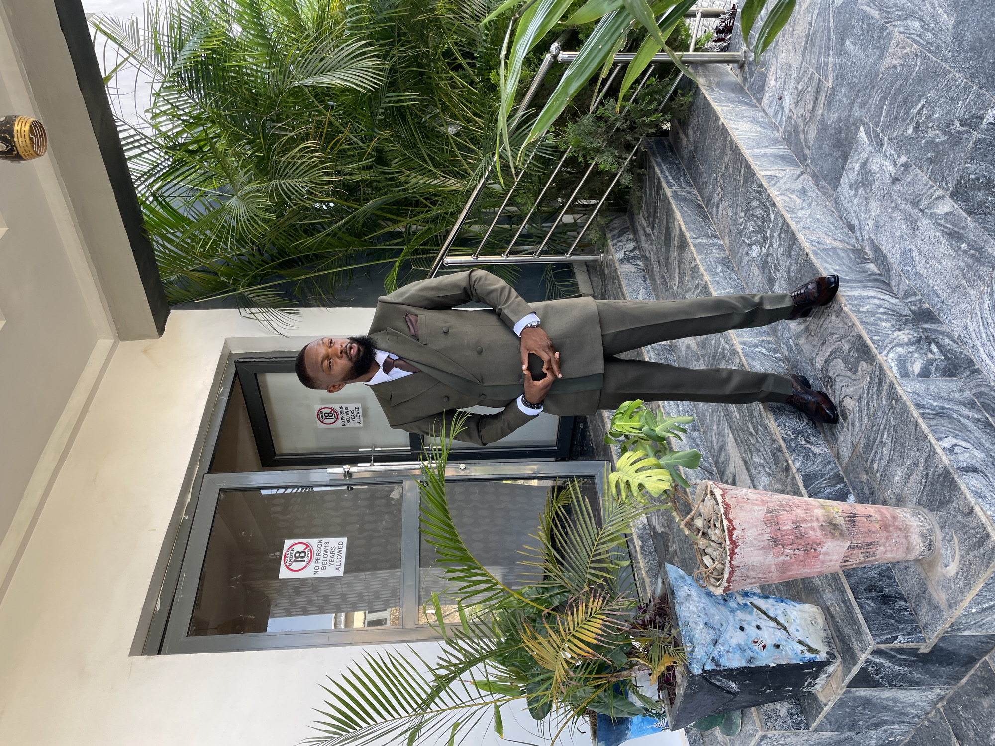 A man in a dark green suit is standing on the steps of a building next to a glass door that has signs posted on it. The signs read, "No Person Below 18 Years Allowed". The man is light-skinned with a beard and is looking at the camera. He is wearing a white dress shirt, brown necktie, a watch on his left wrist, and brown shoes. Behind the man is a wall covered with plants, a stainless steel railing, and a gray marble staircase. A large flower pot filled with plants is at the base of the staircase on the right. The location appears to be Jos, Nigeria based on cultural context and clues in the image.