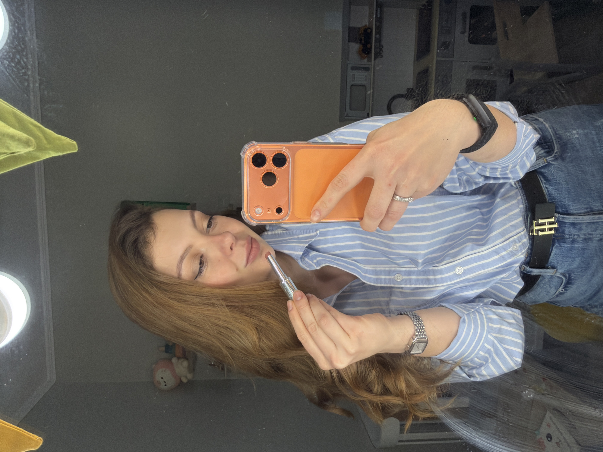 A woman takes a selfie in a mirror. She holds an orange phone in her right hand and applies lipstick with her left. She wears a light blue striped shirt, jeans, a black watch on her left wrist, and a fitness tracker on her right. Her brown hair is shoulder-length. The background shows a room with a light and a plush toy on a shelf, along with other items in the reflection. The location may be Khimki, Russia.