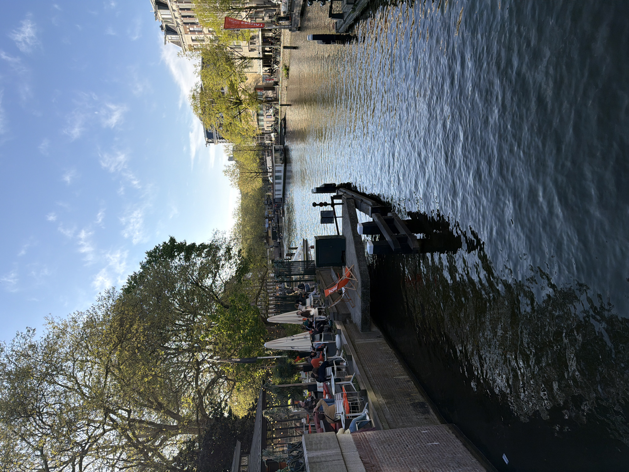 This image depicts a lively canal-side scene in Amsterdam, likely during the late afternoon or early evening on a clear, sunny day. The perspective looks down a canal, with buildings and trees lining both banks.
On the left side of the frame, an outdoor cafe or bar is bustling with activity. Several people are seated at tables under white umbrellas, enjoying the pleasant weather. They appear to be socializing, relaxing, and potentially eating or drinking. The facade of the building housing the cafe displays the text "REZ BAR". Lush green trees with full foliage provide additional shade and frame this side of the canal.
The canal itself dominates the center and right portions of the image, with calm, dark water showing numerous ripples that catch the sunlight. Several houseboats or barges are moored along the banks. In the foreground, a sturdy dark wooden structure, possibly a pier or part of a lock, extends into the water.
The opposite bank features classic Amsterdam architecture: tall, narrow buildings with distinctive gabled roofs. A prominent red banner, partially visible, hangs between two buildings and displays the text "PRINS HENDR", likely referring to Prins Hendrikkade. More trees, some with emerging green leaves and others still with bare branches, line this side of the waterway.
The sky above is bright blue with scattered wispy white clouds, indicating excellent weather. The lighting casts soft shadows and highlights on the water, contributing to a relaxed and inviting atmosphere. The overall scene captures the typical charm of Amsterdam's canals and outdoor urban life.