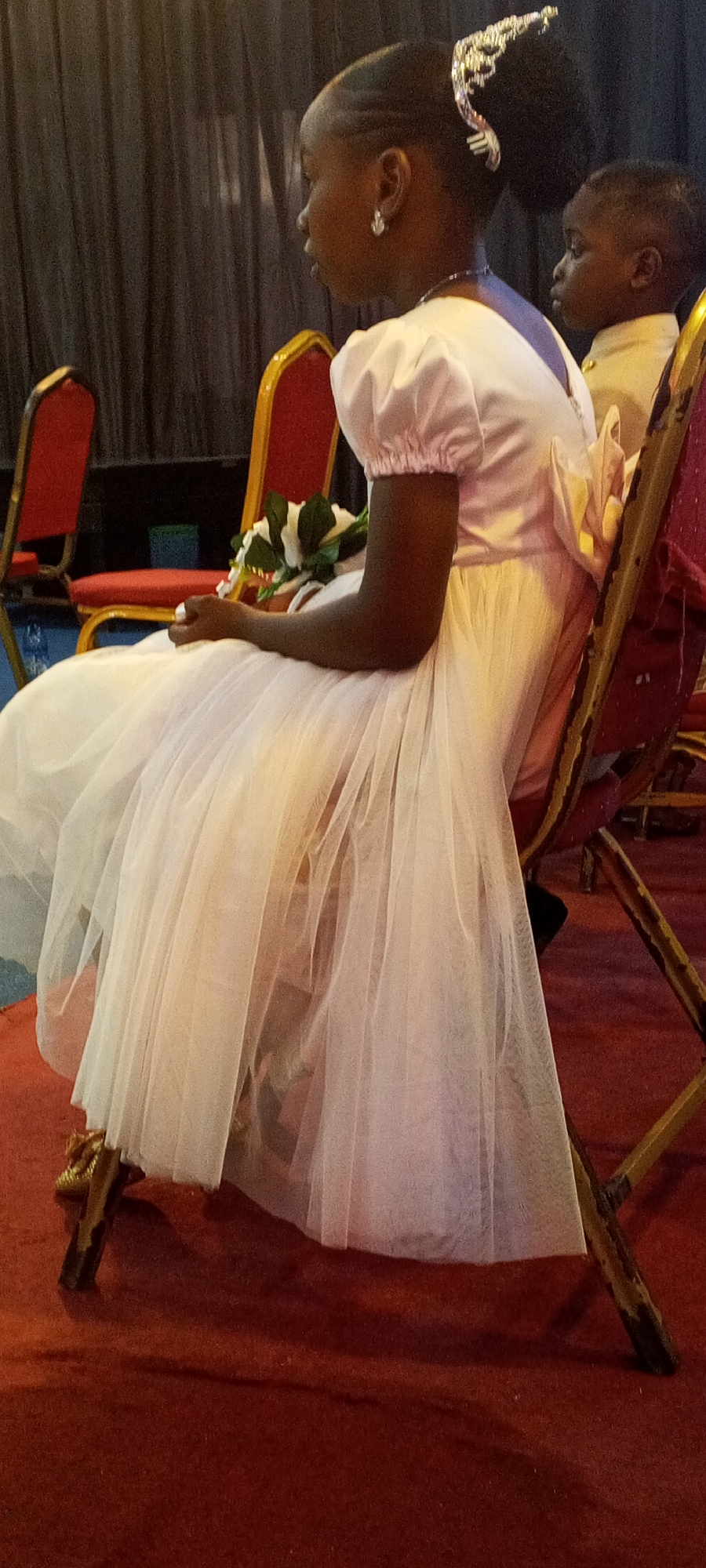A young, dark-skinned girl with braided hair adorned with a jeweled hairpiece, sits on a gold-colored chair. She is dressed in a light pink dress with a puffy skirt. She is holding a bouquet of artificial white and green flowers. To her right is another dark-skinned child wearing a light yellow shirt. The setting appears to be indoors, with a red carpet and dark curtains. The photo was taken in the city of Sabon Tasha, Nigeria.