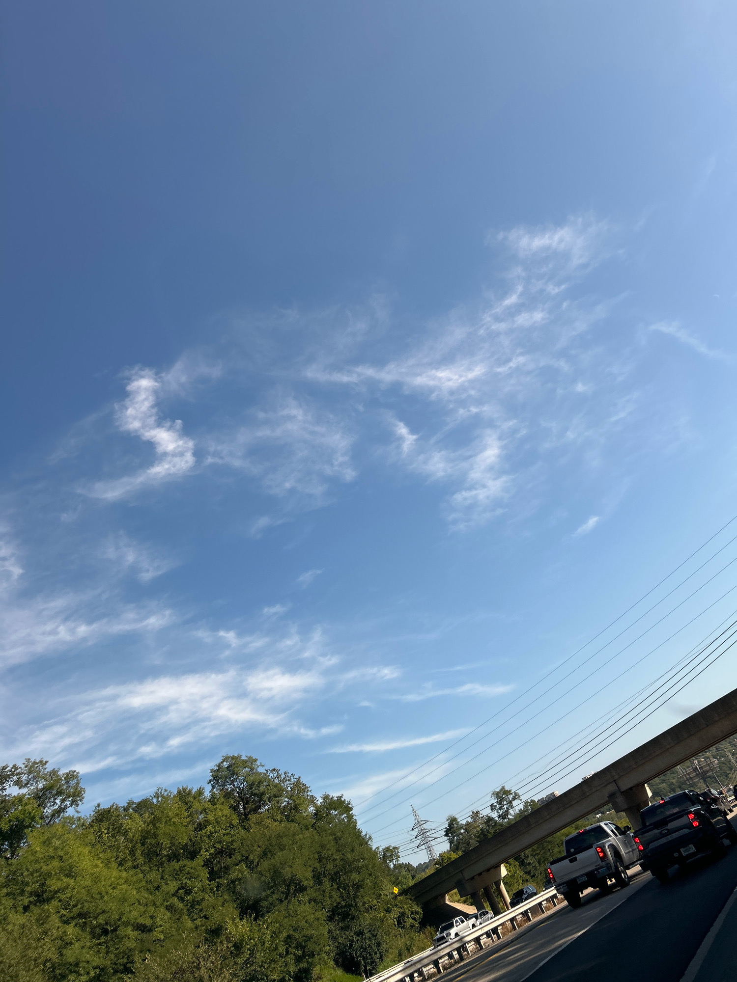 A bright, sunny day is captured from a low angle looking up at a vast blue sky with scattered wispy white clouds. In the foreground, a road is visible with several vehicles, including trucks and cars, driving on it. A concrete overpass looms overhead, supported by thick pillars, and power lines stretch across the scene. To the left, a dense line of green trees forms a natural barrier. The location is identified as Collier Township, United States of America.